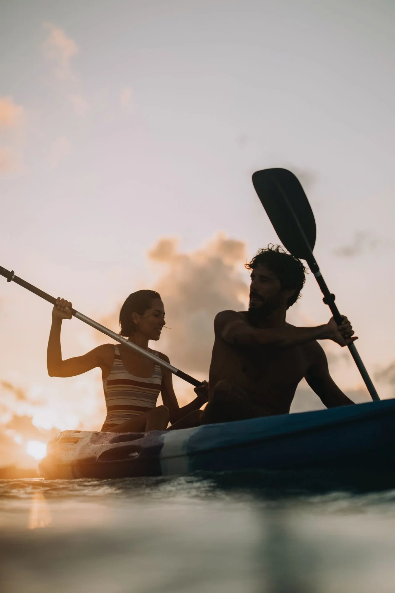 A silhouettes of a man and woman kayaking on the water during sunset or sunrise, with clouds in the sky.