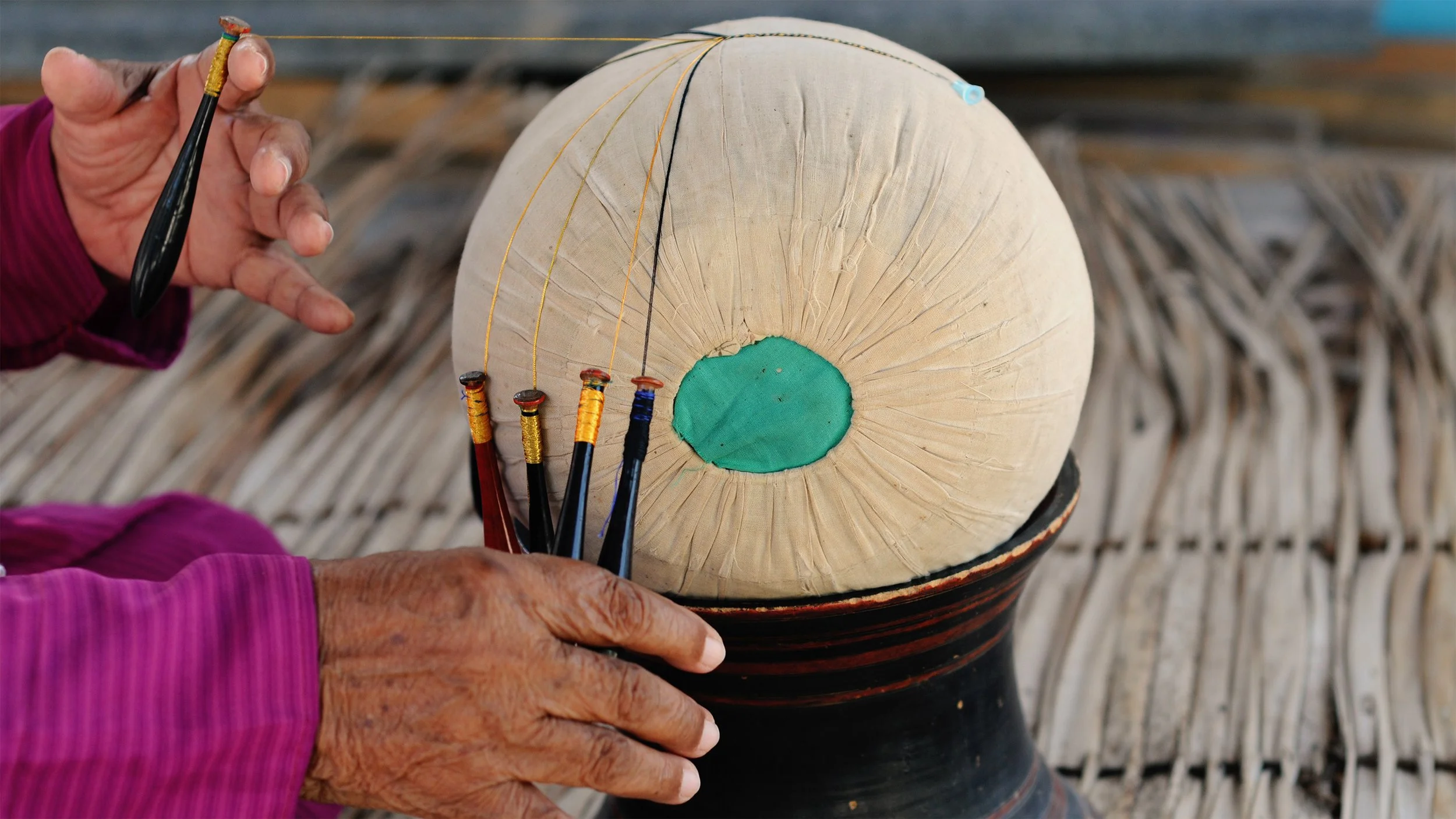 Close-up of an older person's hands making a traditional drum with a round, drumhead and colorful decorative elements, with fishing lines or strings attached.