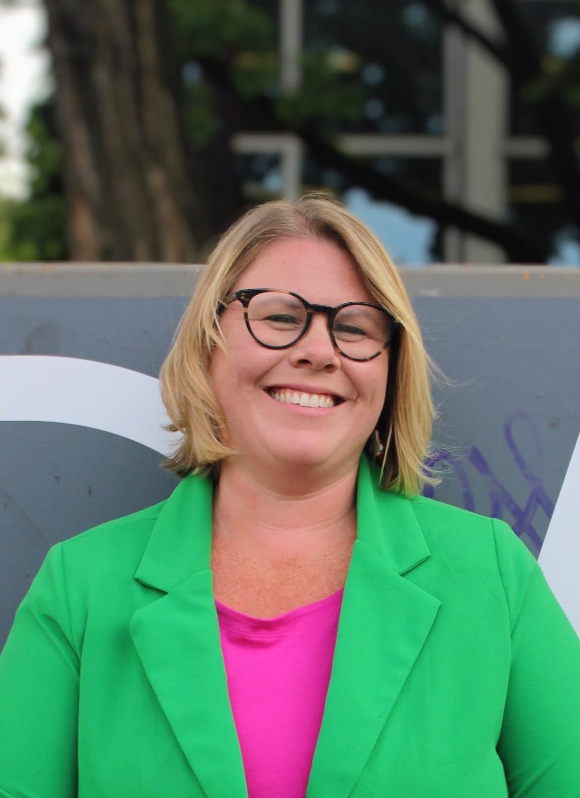 A woman with blonde hair, wearing glasses, a green blazer, and a pink top, smiling outdoors in front of a sign.