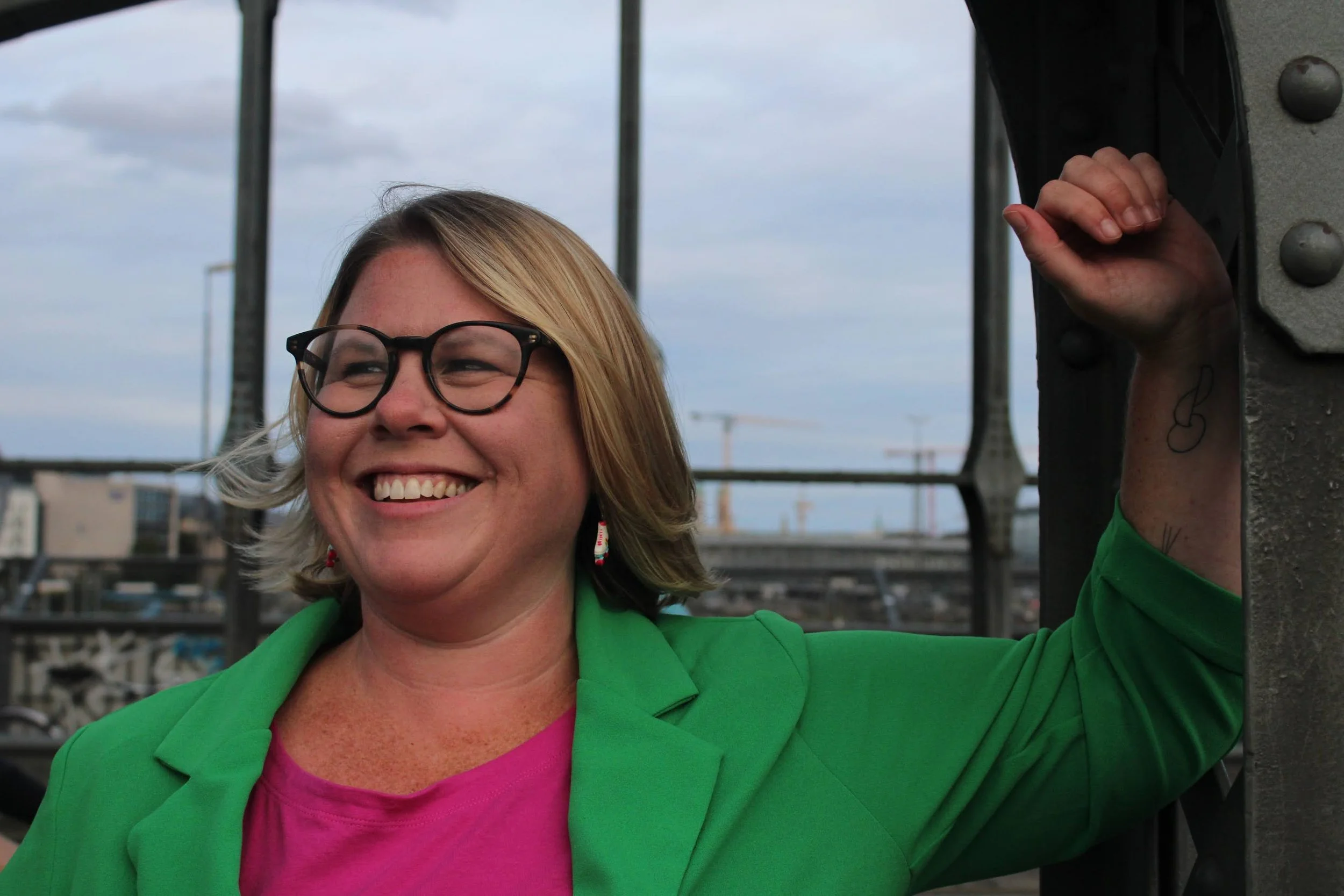 A woman with blonde hair, glasses, and a bright smile, wearing a green blazer over a pink top, standing outdoors with a metal structure and a cityscape in the background.