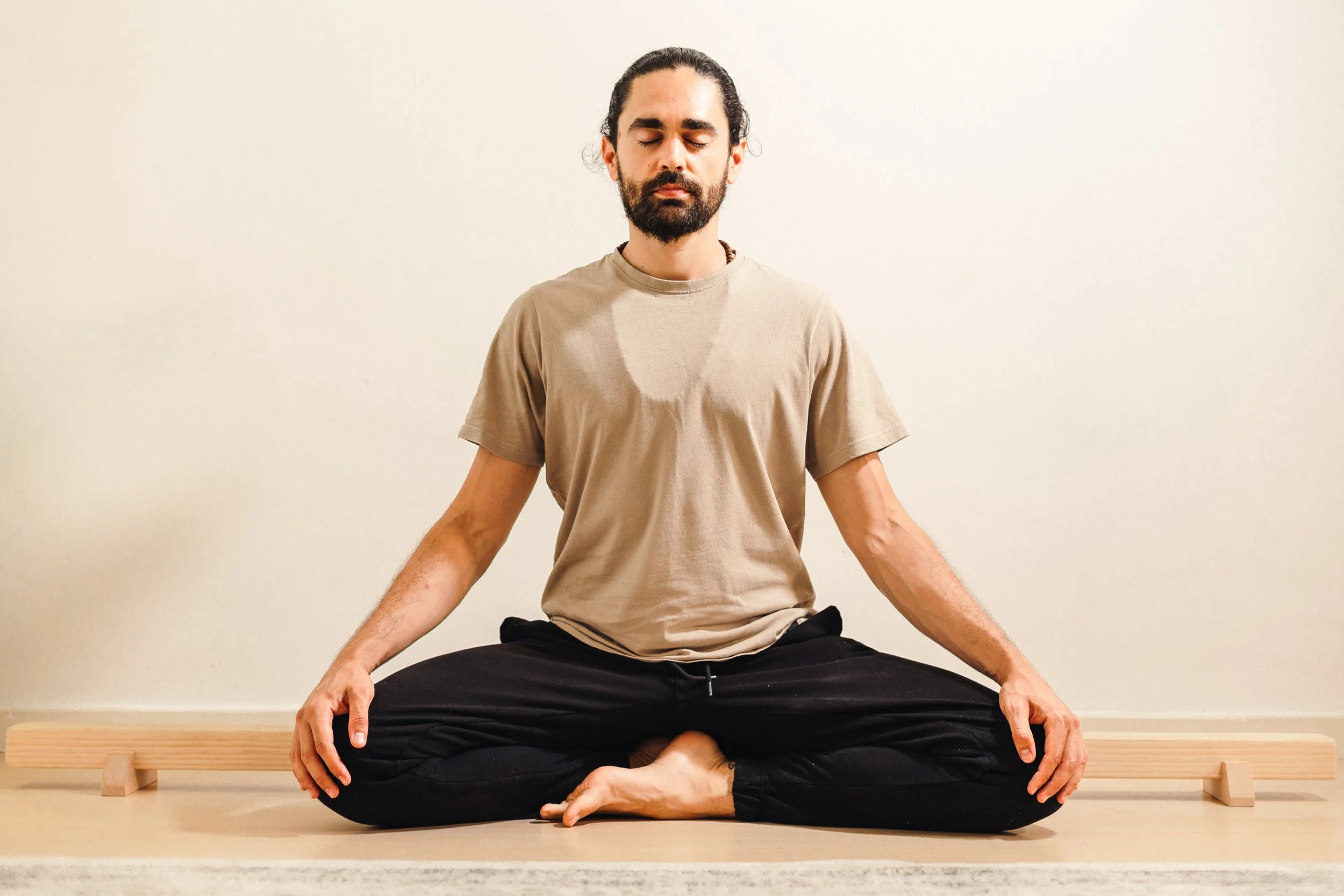 Un hombre con barba y cabello recogido en una coleta, sentado en posición de meditación con las piernas cruzadas en un suelo de madera, con una pared blanca de fondo, ojos cerrados y expresión tranquila.