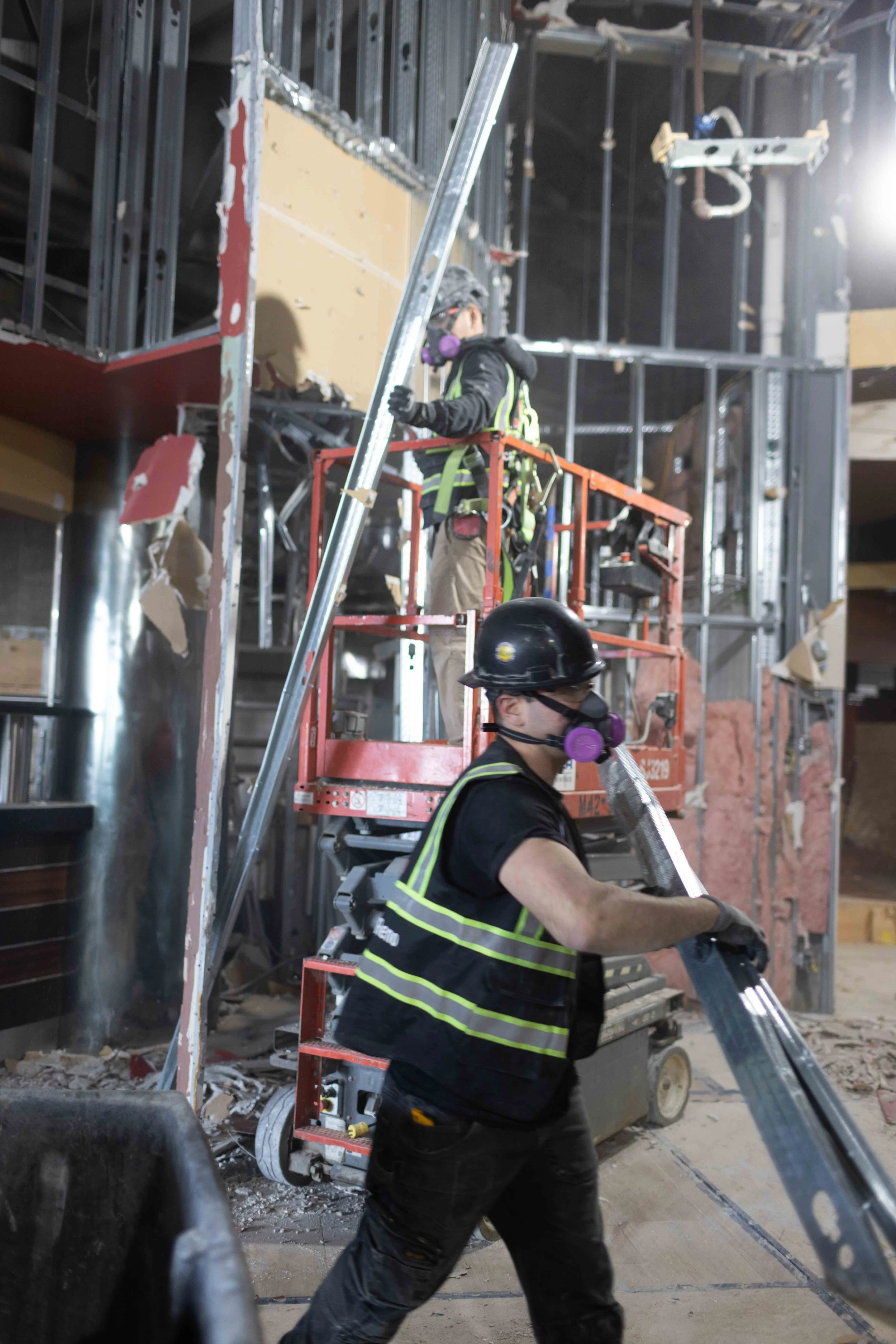 Construction workers in safety gear working on a building site, using lifts and tools amidst a partially constructed interior with exposed framing and debris on the floor.
