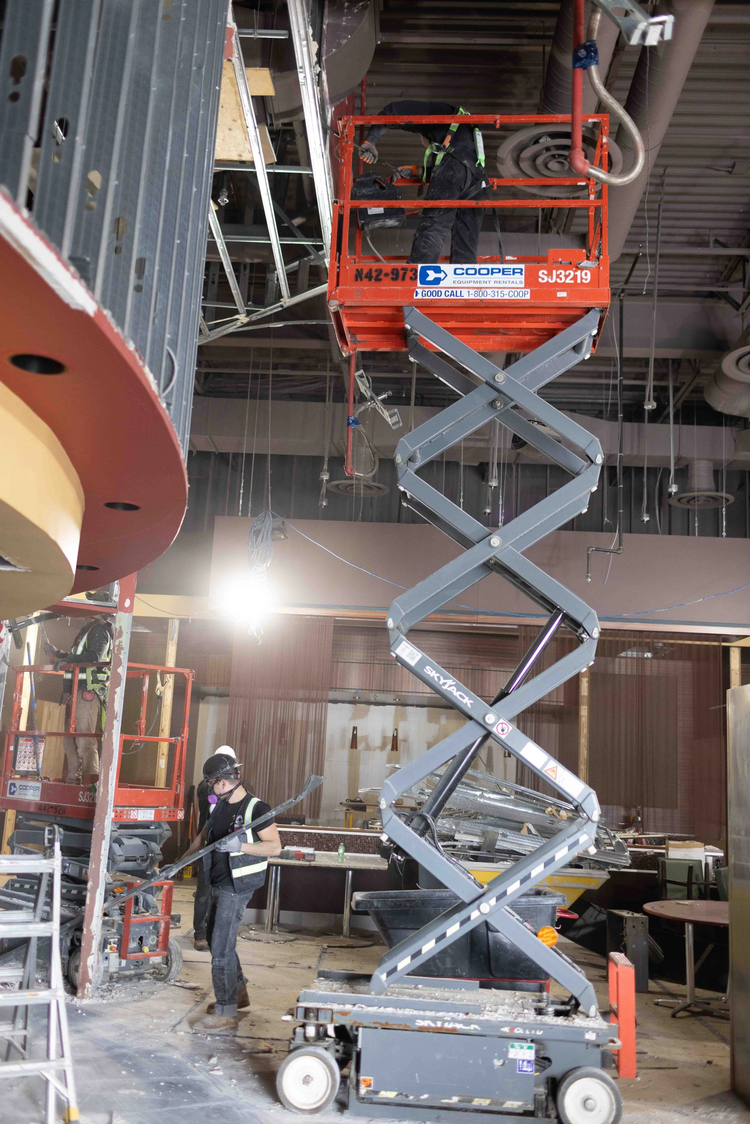 Workers in safety gear using cherry picker and scaffolding to perform construction or renovation work inside a building, with various tools and equipment around.