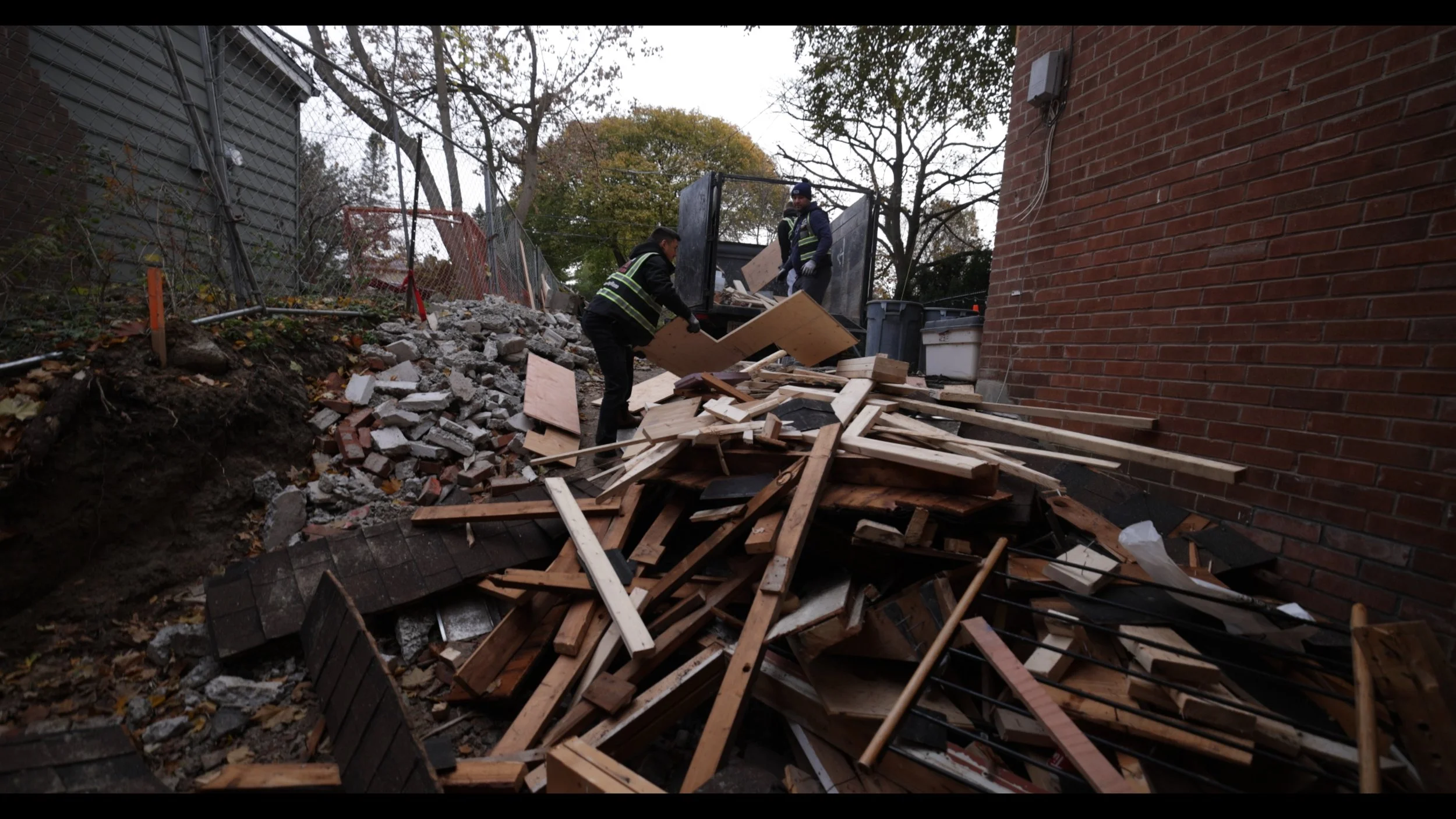 Two workers in reflective vests clearing debris, including wood and rubble, from a collapsed area between a brick wall and a fence.