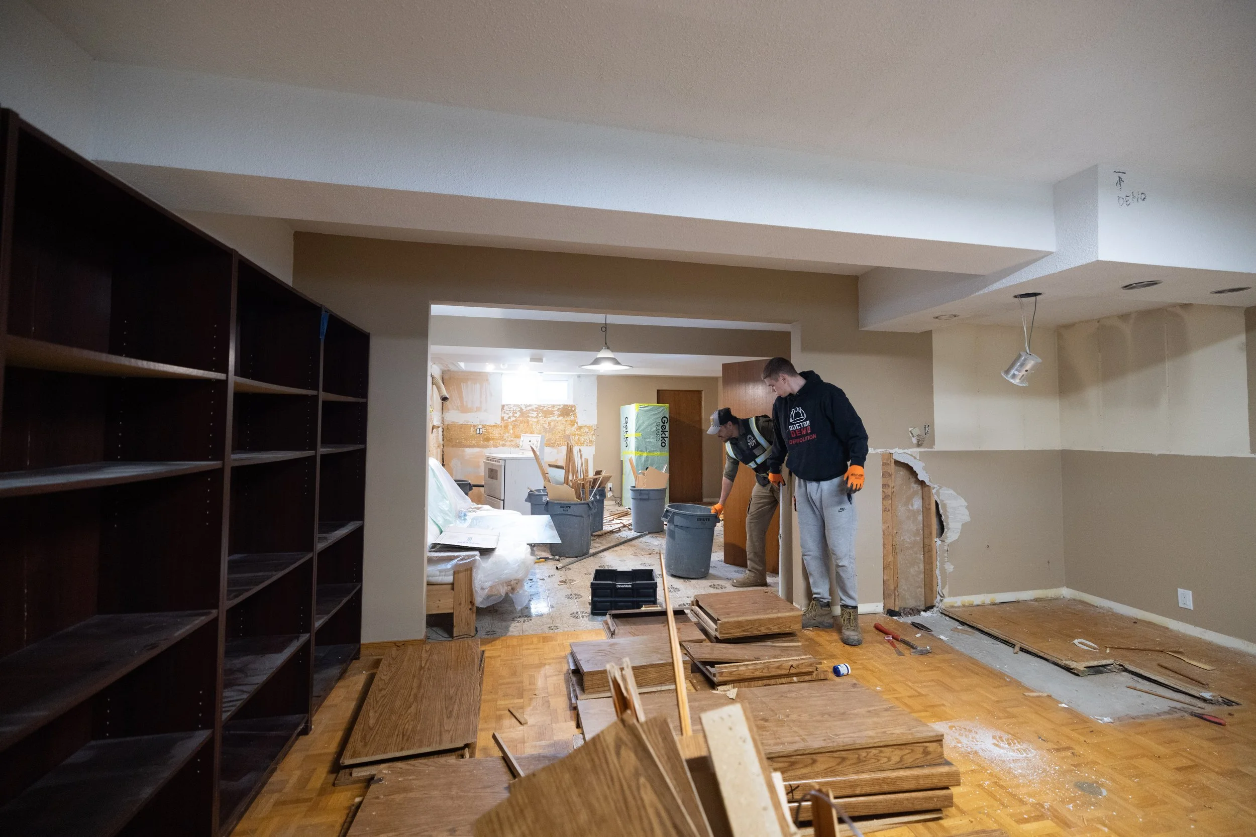 Two workers are remodeling a room with exposed walls and scattered wooden flooring materials and tools around them.