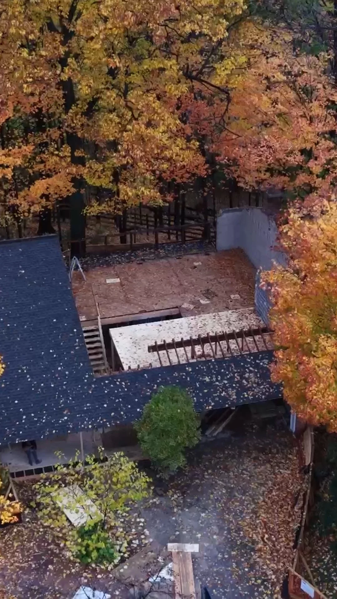An aerial view of a house with a partially demolished backyard and colorful fall trees surrounding it.