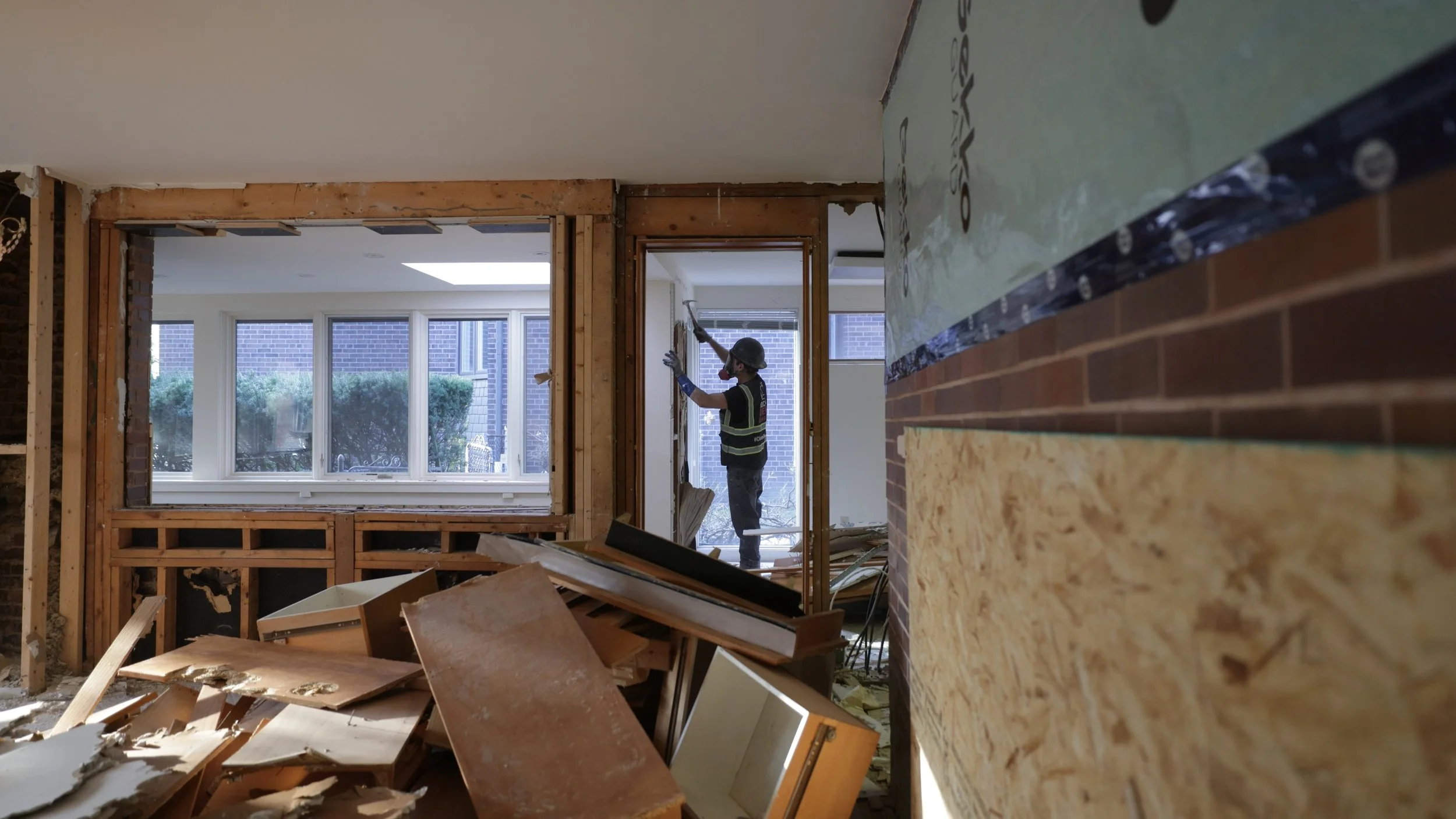 A construction worker wearing a helmet and safety vest is working inside a house under renovation, using a tool on the wall near a window with brick exterior visible outside.