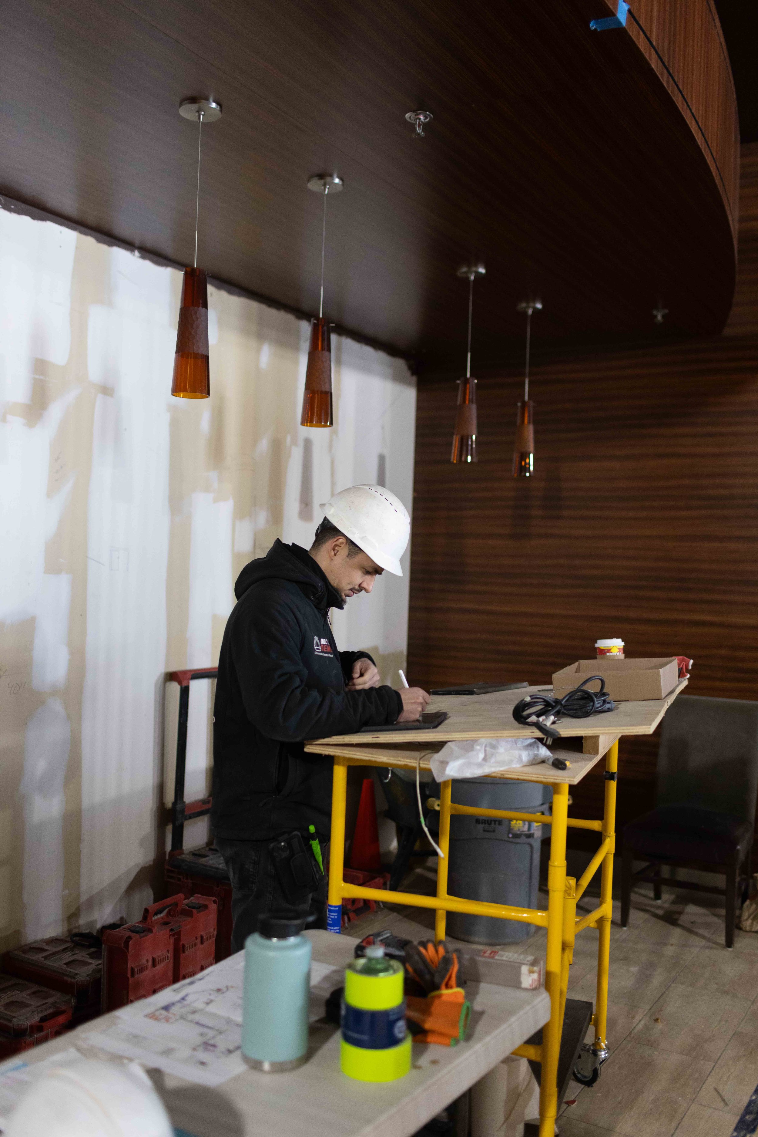 A construction worker wearing a white safety helmet and dark jacket is standing at a work table, working on a project. The table is cluttered with various tools and materials, including bottles, tape, and gloves. The environment appears to be indoors