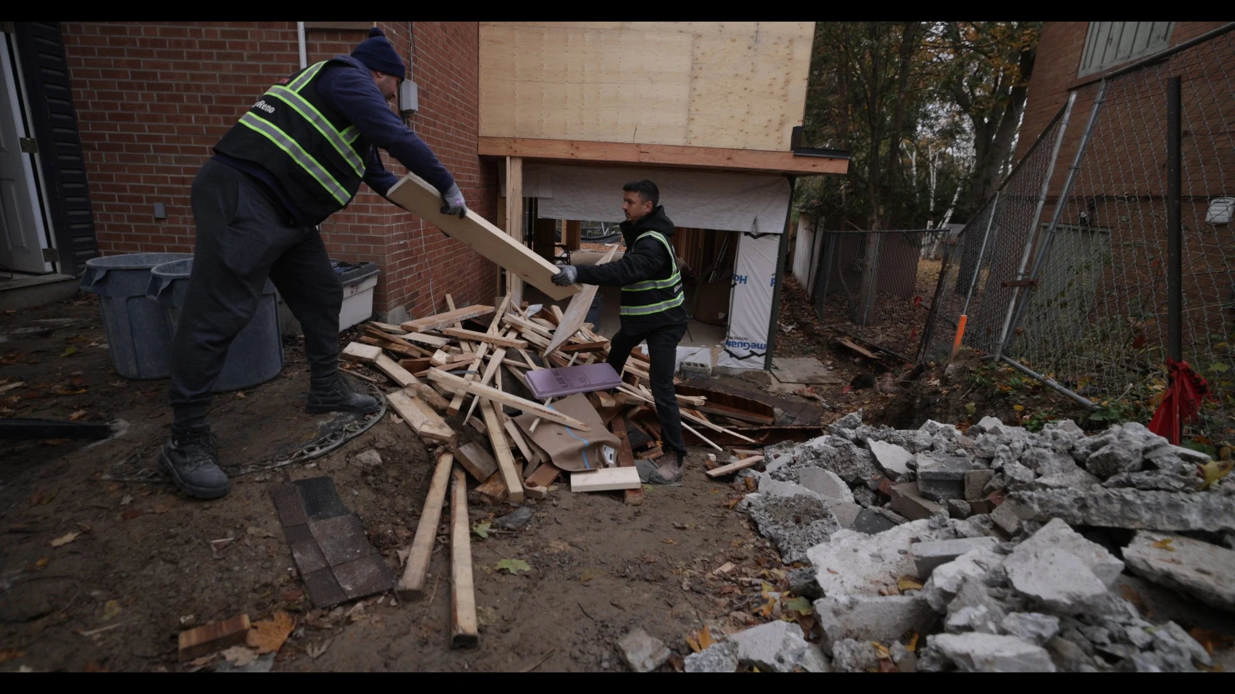 Two workers in safety vests and gloves are clearing debris from a construction or demolition site next to a brick building, with a pile of wood and rubble on the ground.