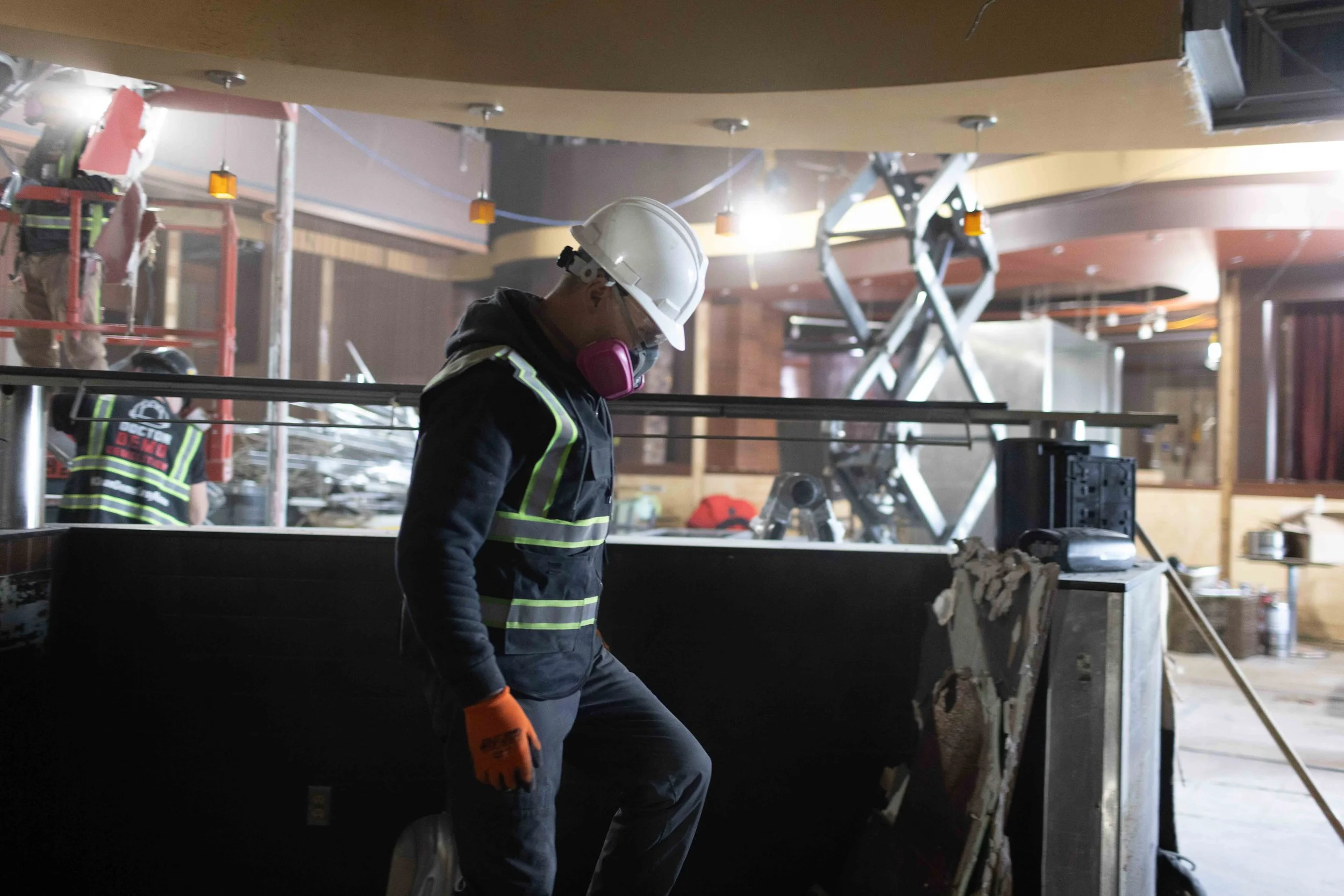 A construction worker in a hard hat and safety gear walking inside a building under construction, with scaffolding and construction equipment visible in the background.