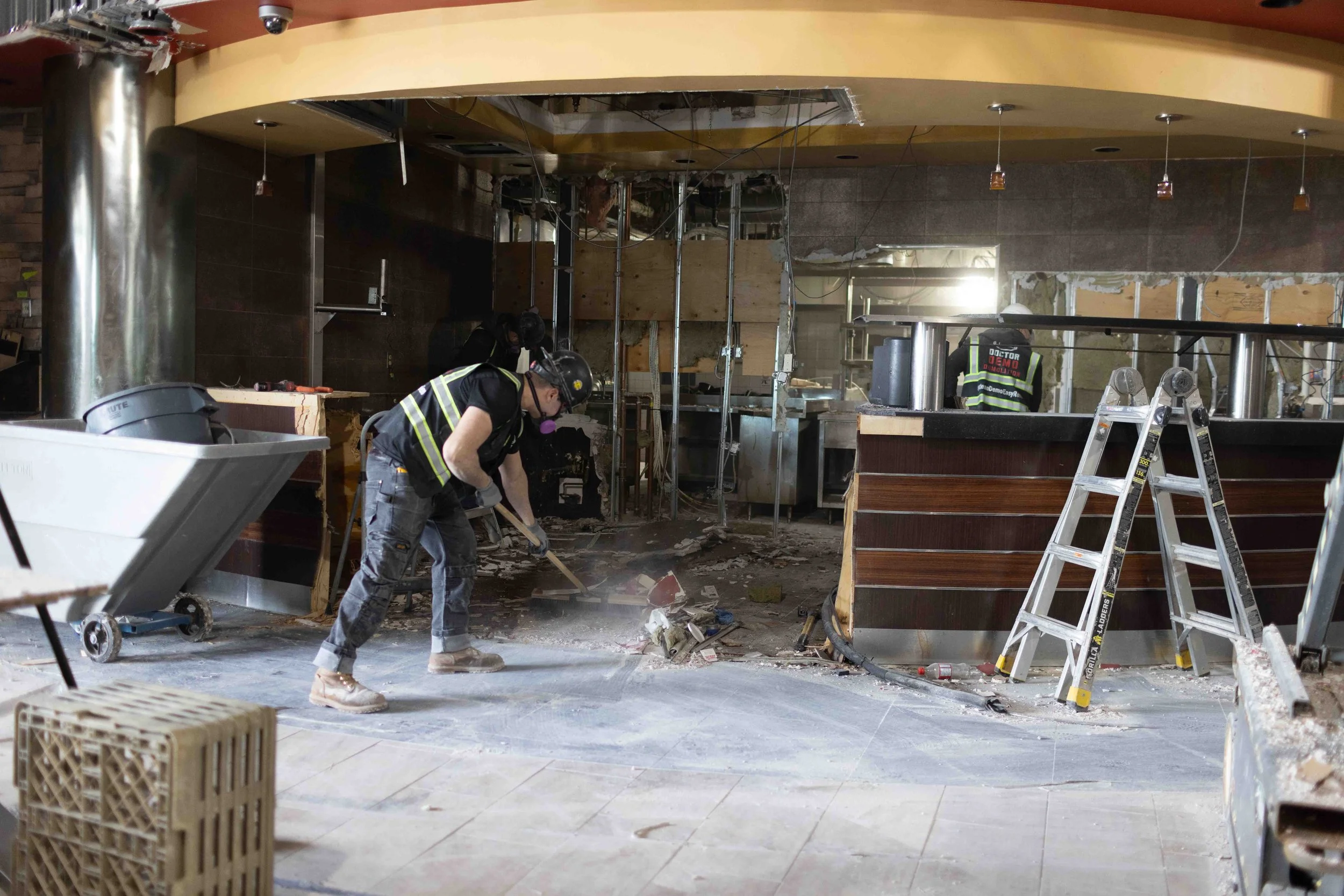 Workers demolishing the interior of a restaurant or store, with debris on the floor, partially dismantled walls, and exposed ceiling fixtures.