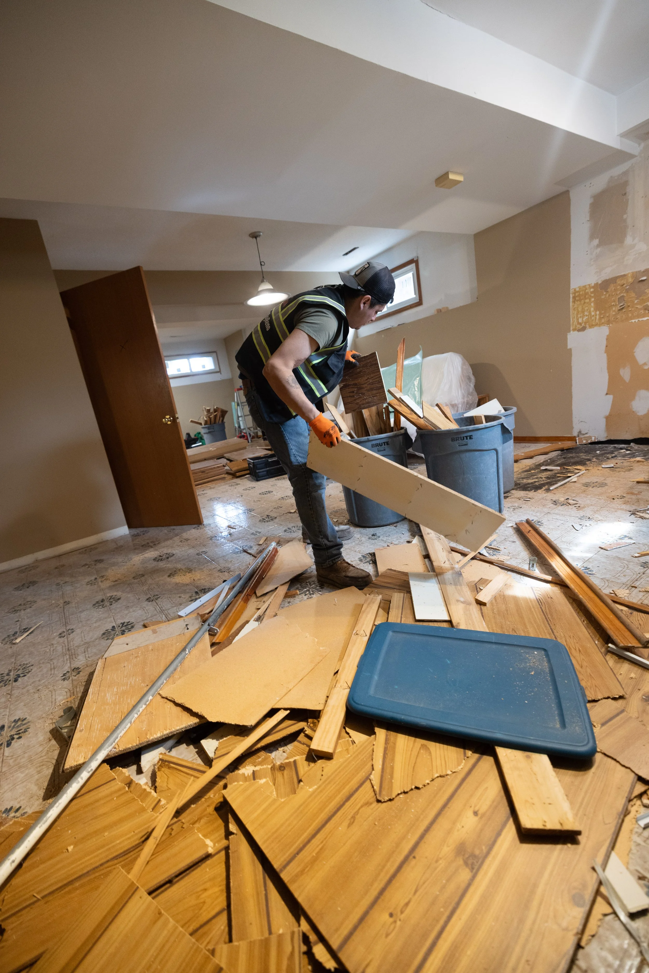 A man wearing a black cap, safety vest, and orange gloves is working in a room under renovation, surrounded by scattered wooden planks, tools, and renovation materials.
