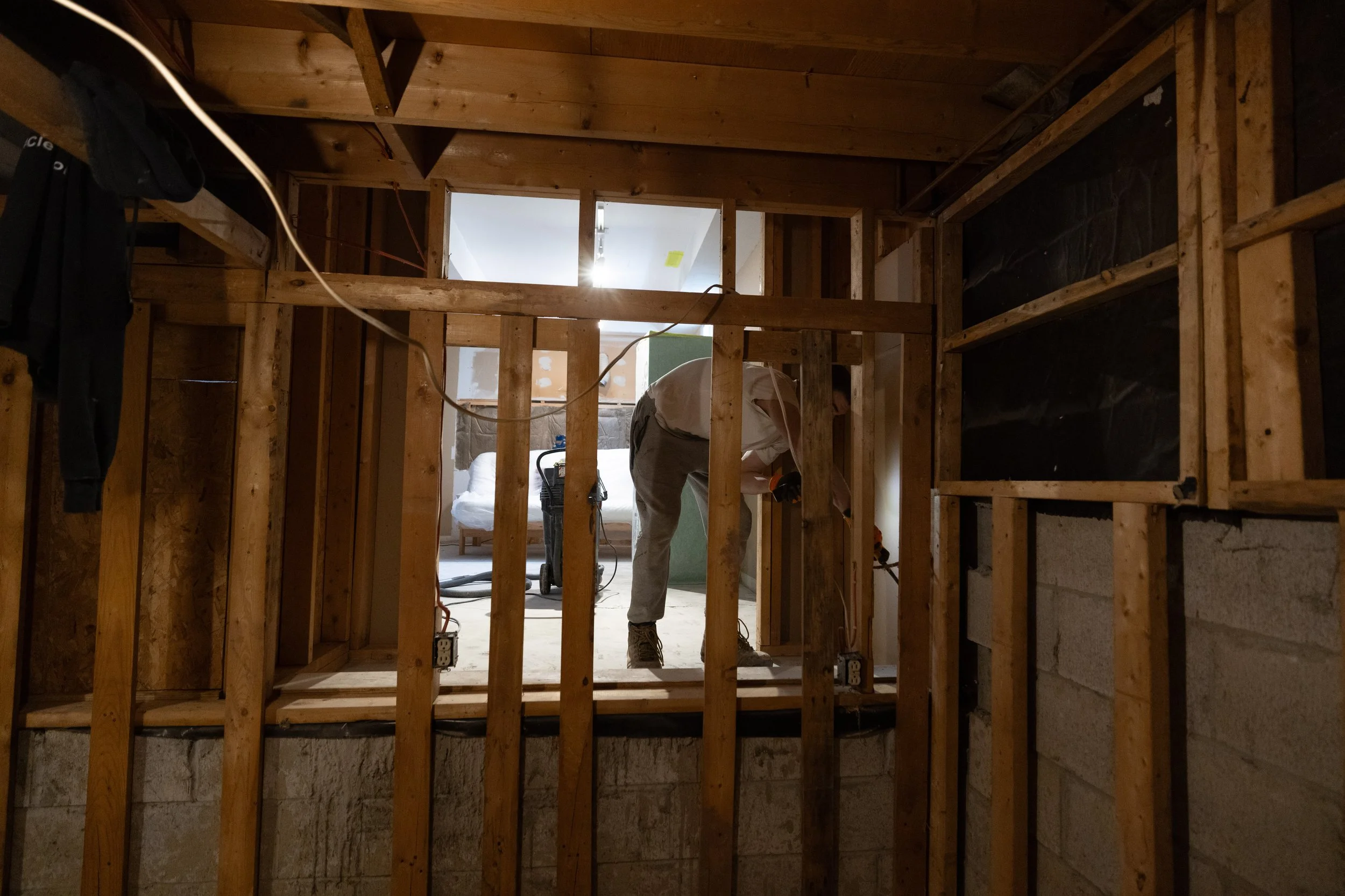 A person working on plumbing inside a partially constructed room with exposed wooden framing and concrete Block walls.