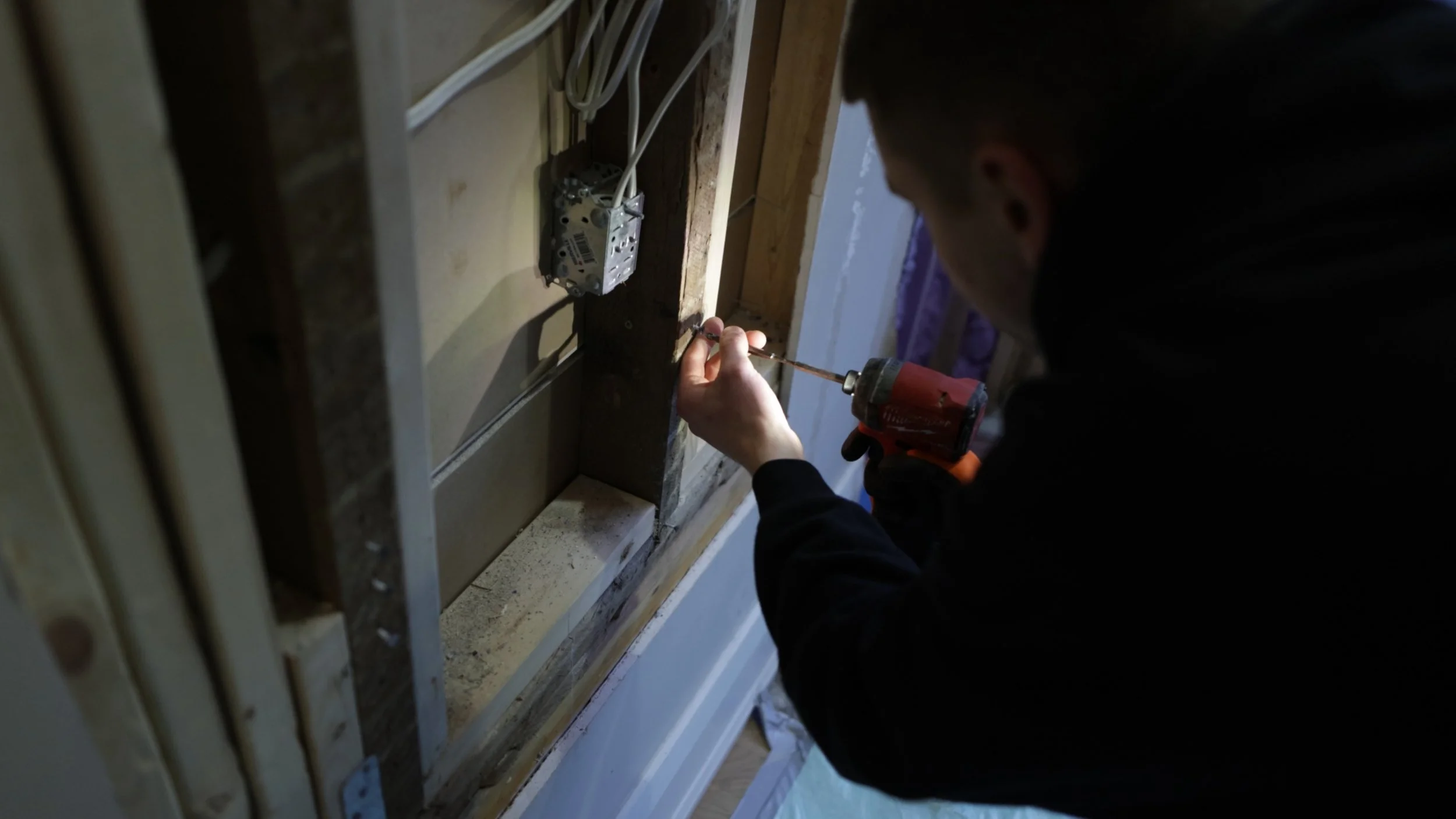 A person working on electrical wiring inside a wall, holding a drill and using a screwdriver near an electrical box.