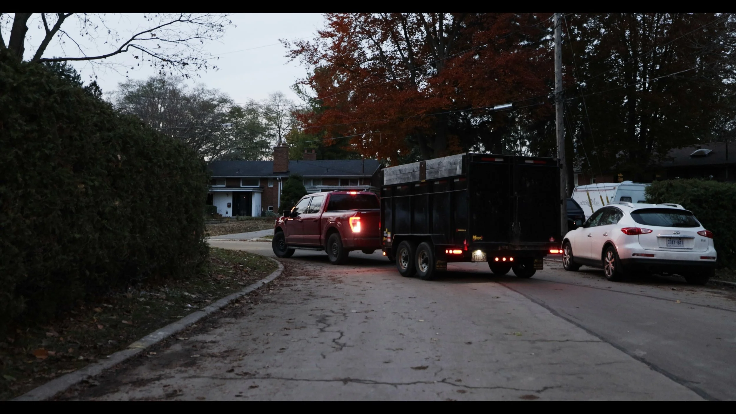 A residential street scene at dusk showing a red truck towing a black trailer, a white car, and a house with a brick chimney, trees with autumn leaves, and utility poles with power lines.