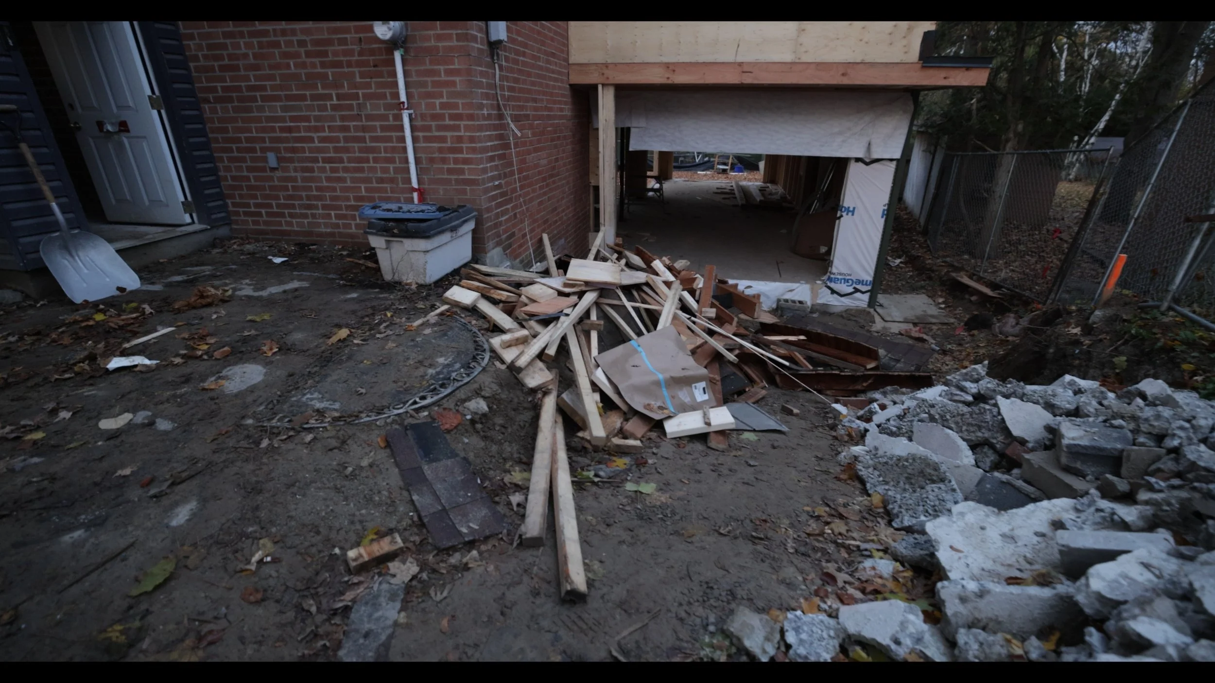 Construction site with wood scraps, debris, and partially built structure next to a brick house and fenced yard.
