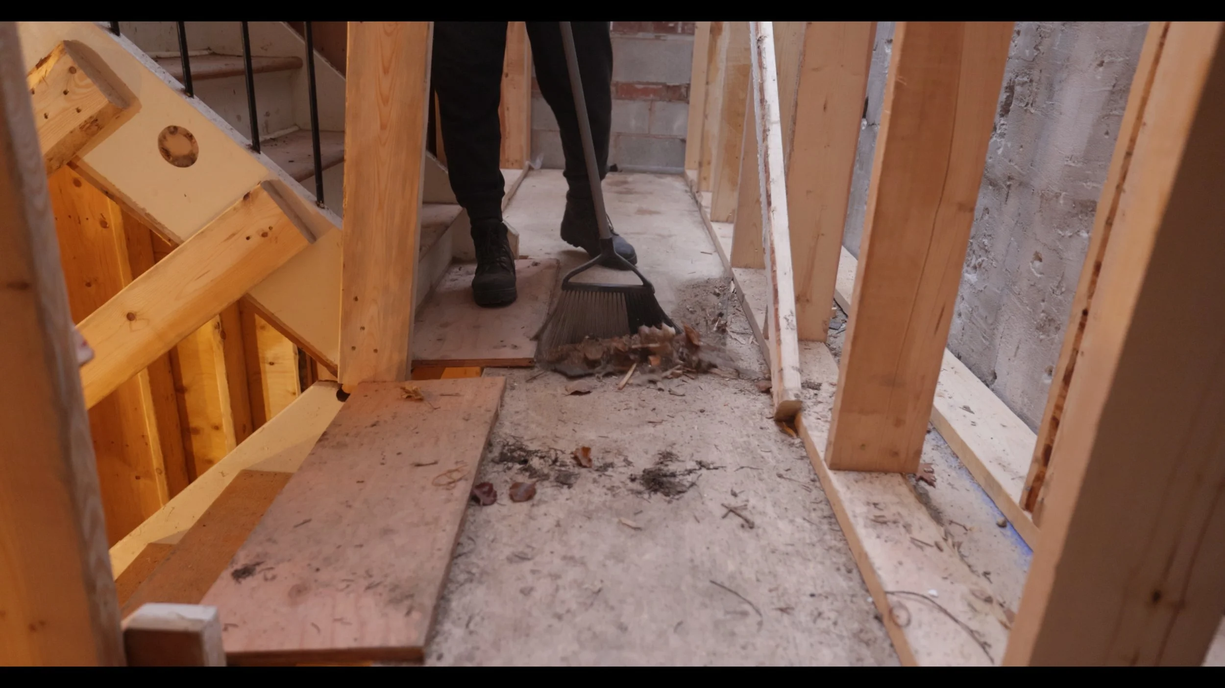 A person sweeping debris from a construction site with exposed wooden framing and concrete flooring.