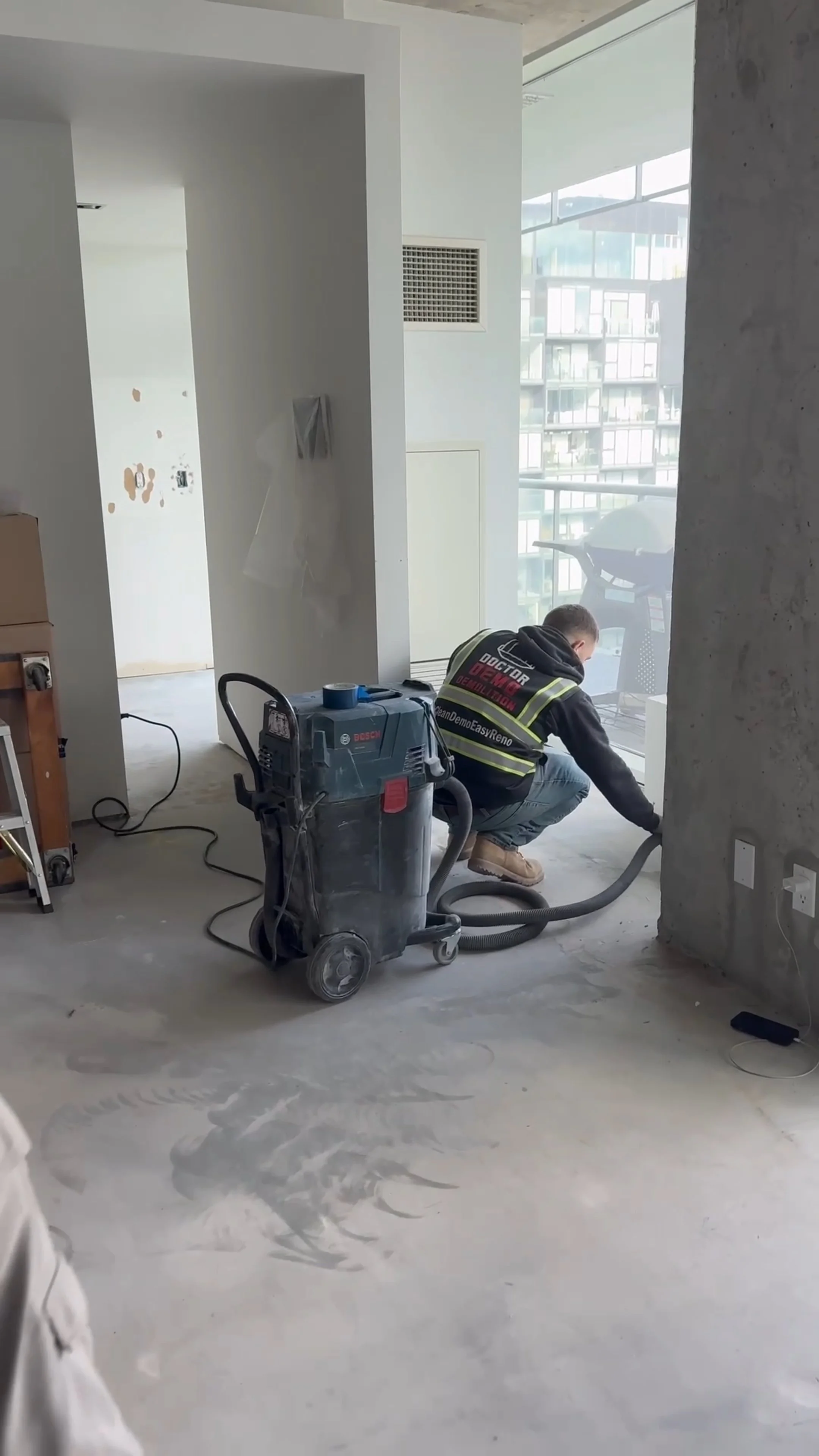 A worker in black jacket and jeans is working on the floor of a modern apartment with large windows. Construction equipment, including a vacuum cleaner, is present in the unfinished space.