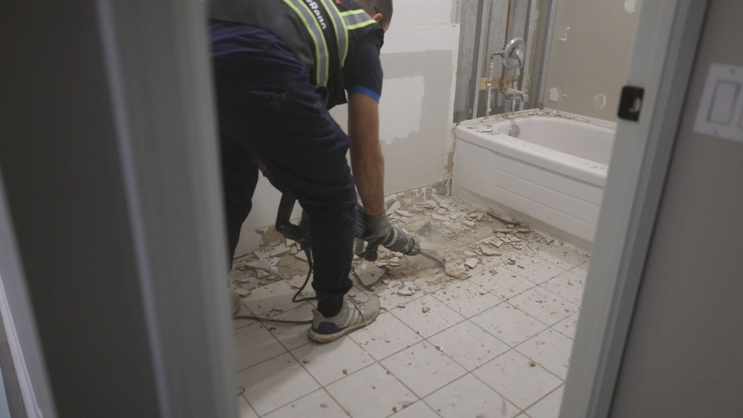 A person using a hammer drill to demolish a tile wall in a bathroom, with debris on the floor and a bathtub visible in the background.