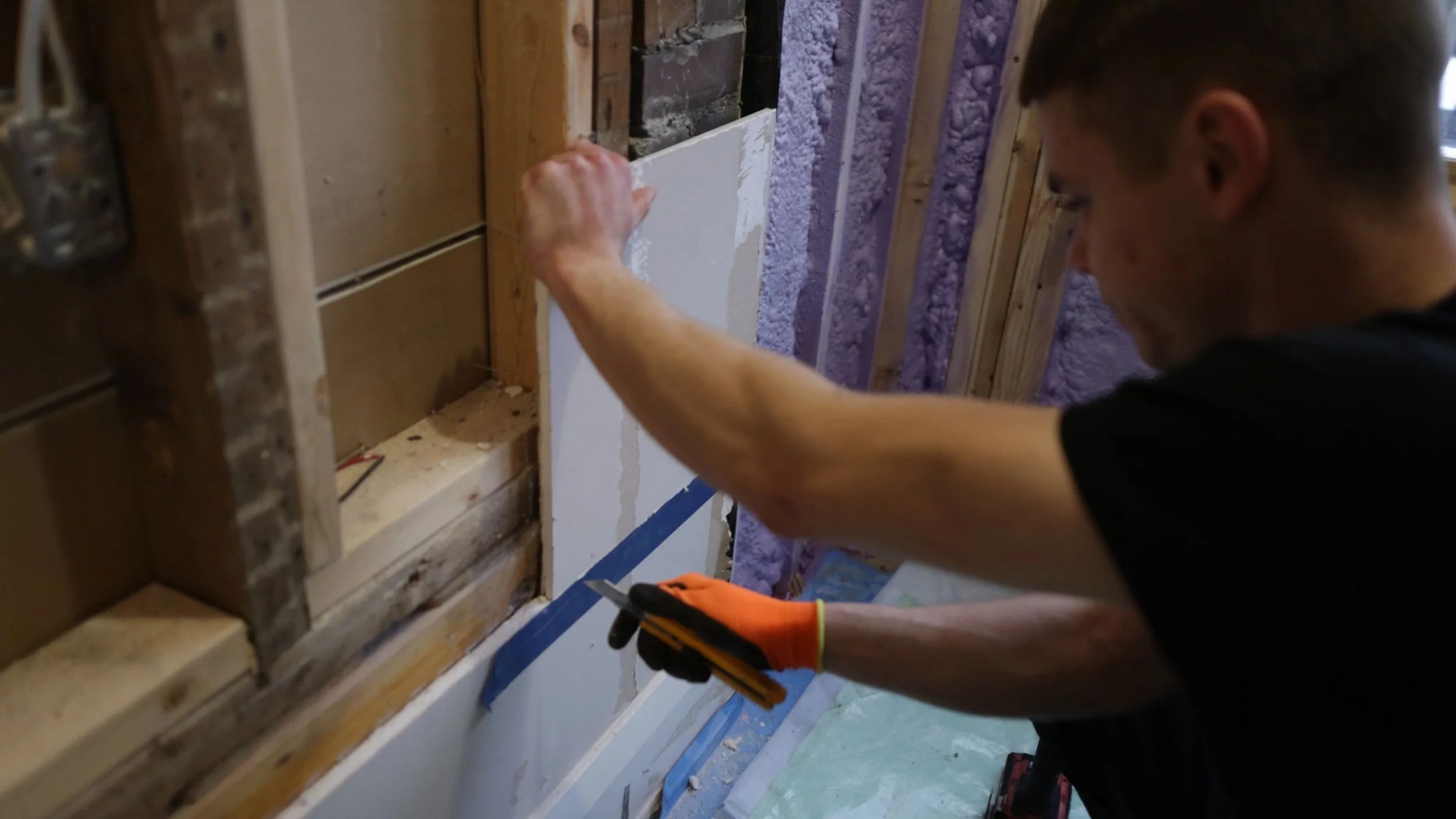 A person working on wiring or electrical work inside a wall with exposed wooden studs and insulation, holding a tool and wearing a glove.