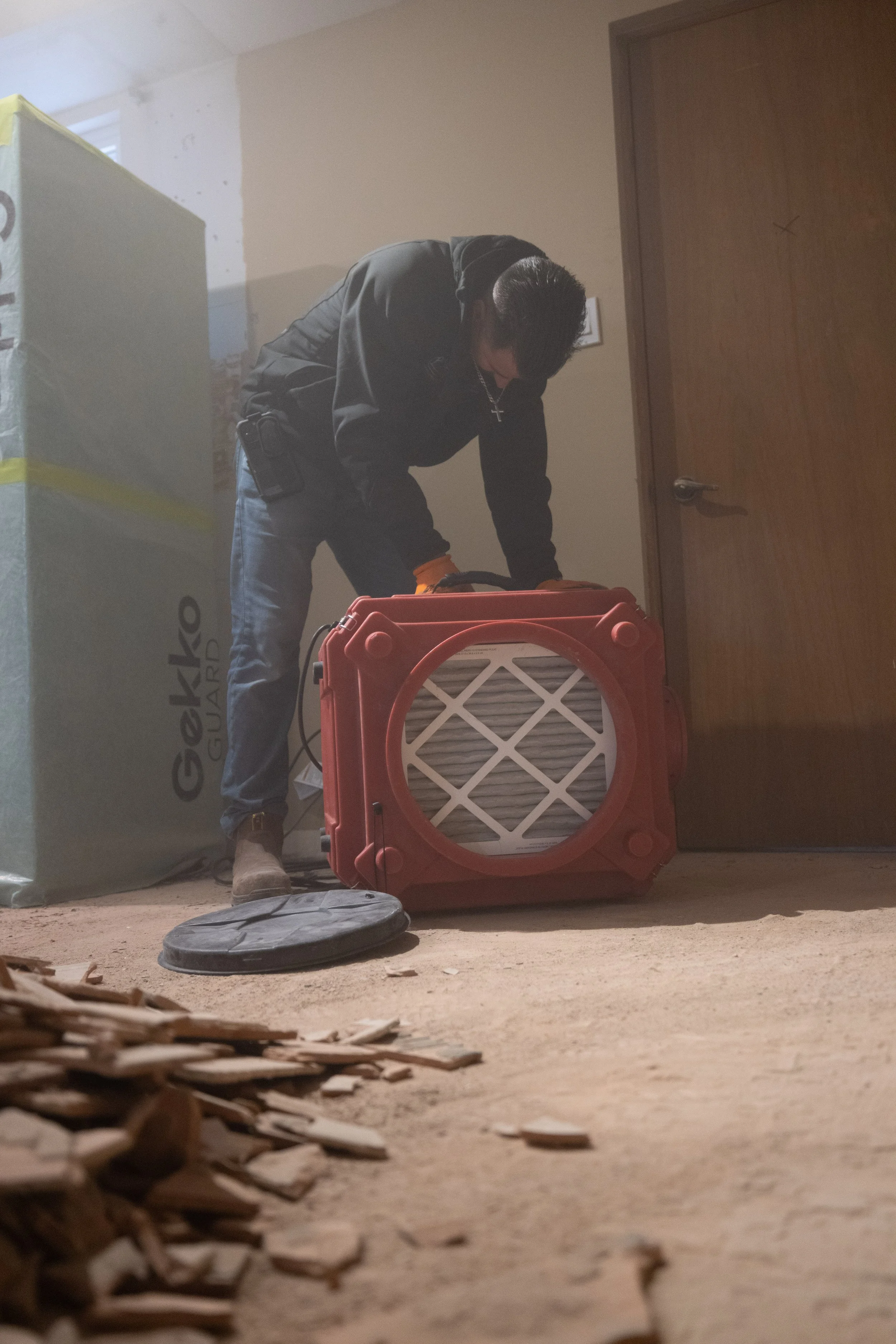 Man working with construction equipment in a room with a cardboard box, a wooden door, and a piece of construction equipment.