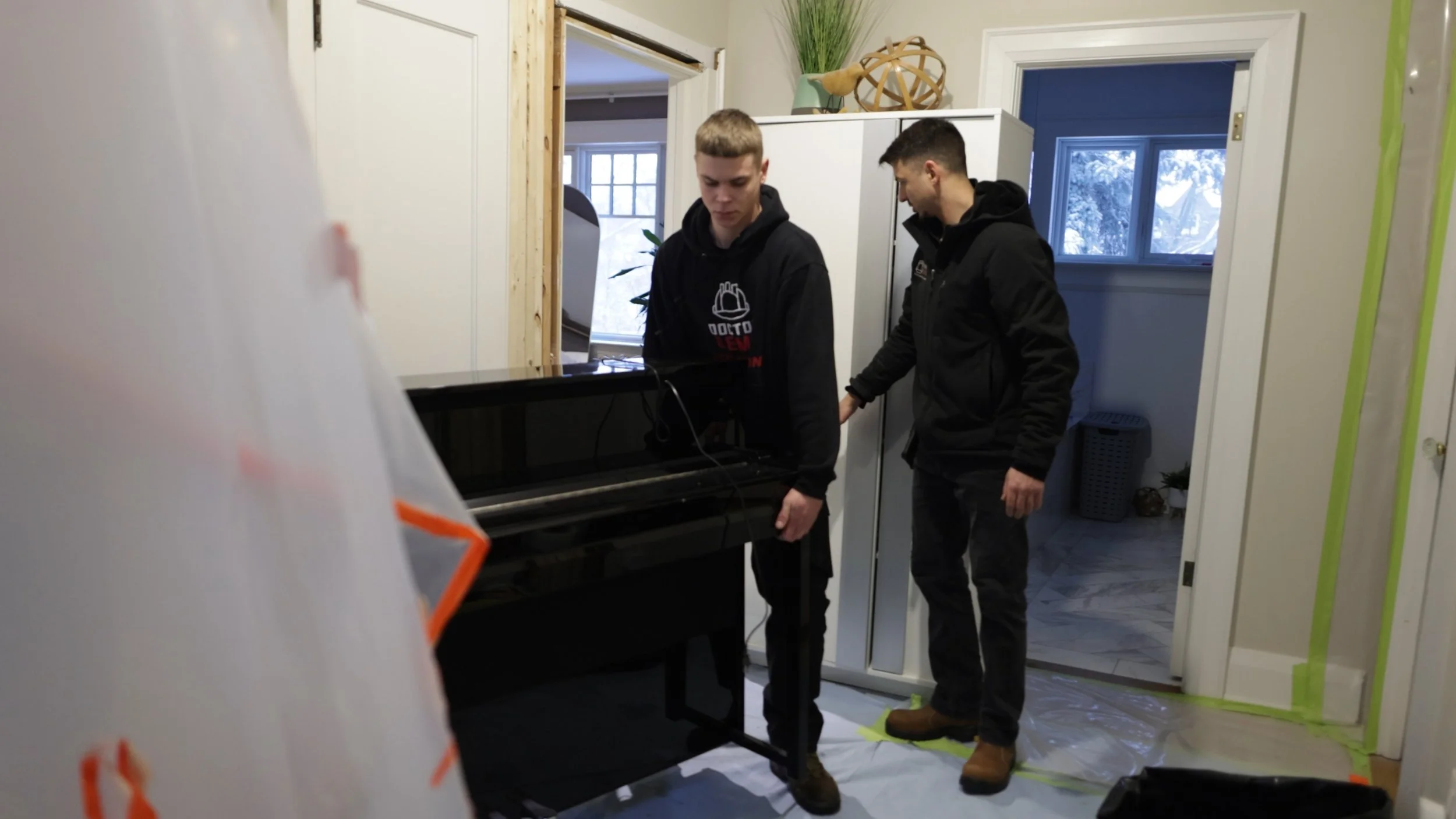 Two young men are working on moving a piano inside a house. They are wearing black hooded jackets and jeans, and are standing near a white cabinet and a window. The room has a light-colored doorframe, green tape on the floor, and some decorative item
