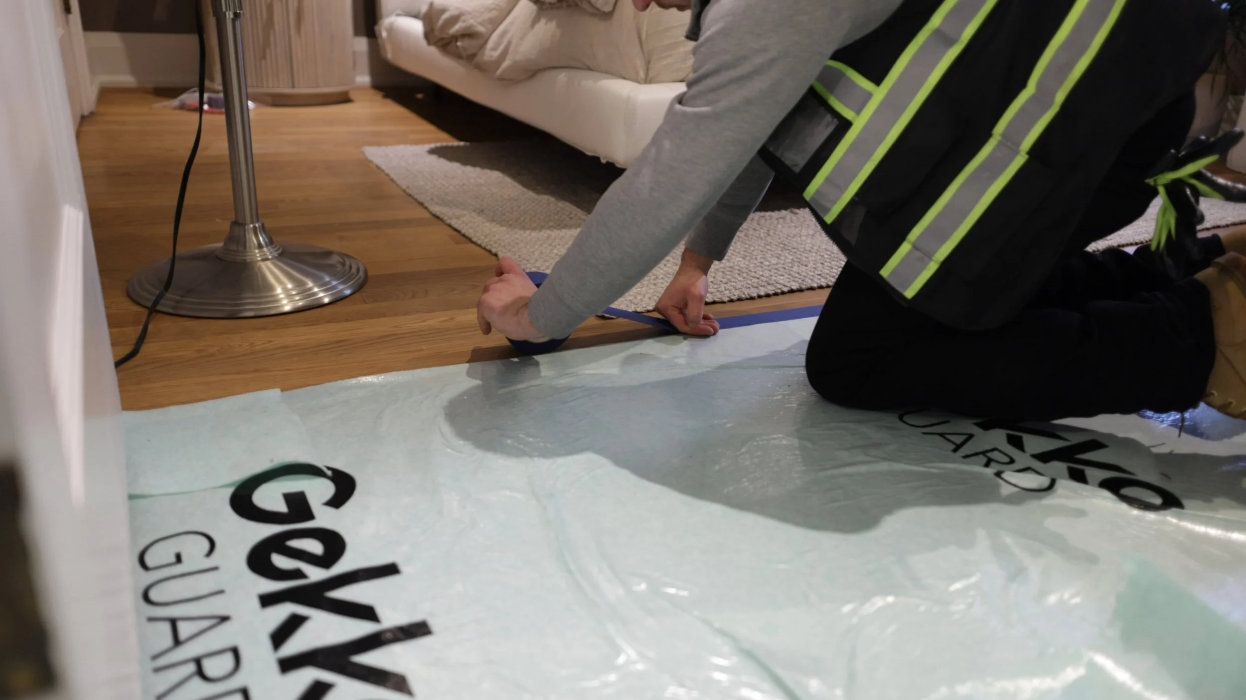 A person kneeling on the floor and installing a plastic sheet under a carpet in a living room. They are wearing a gray shirt, black pants with reflective stripes, and work boots. There is a sofa, a lamp, and a wooden floor in the room.
