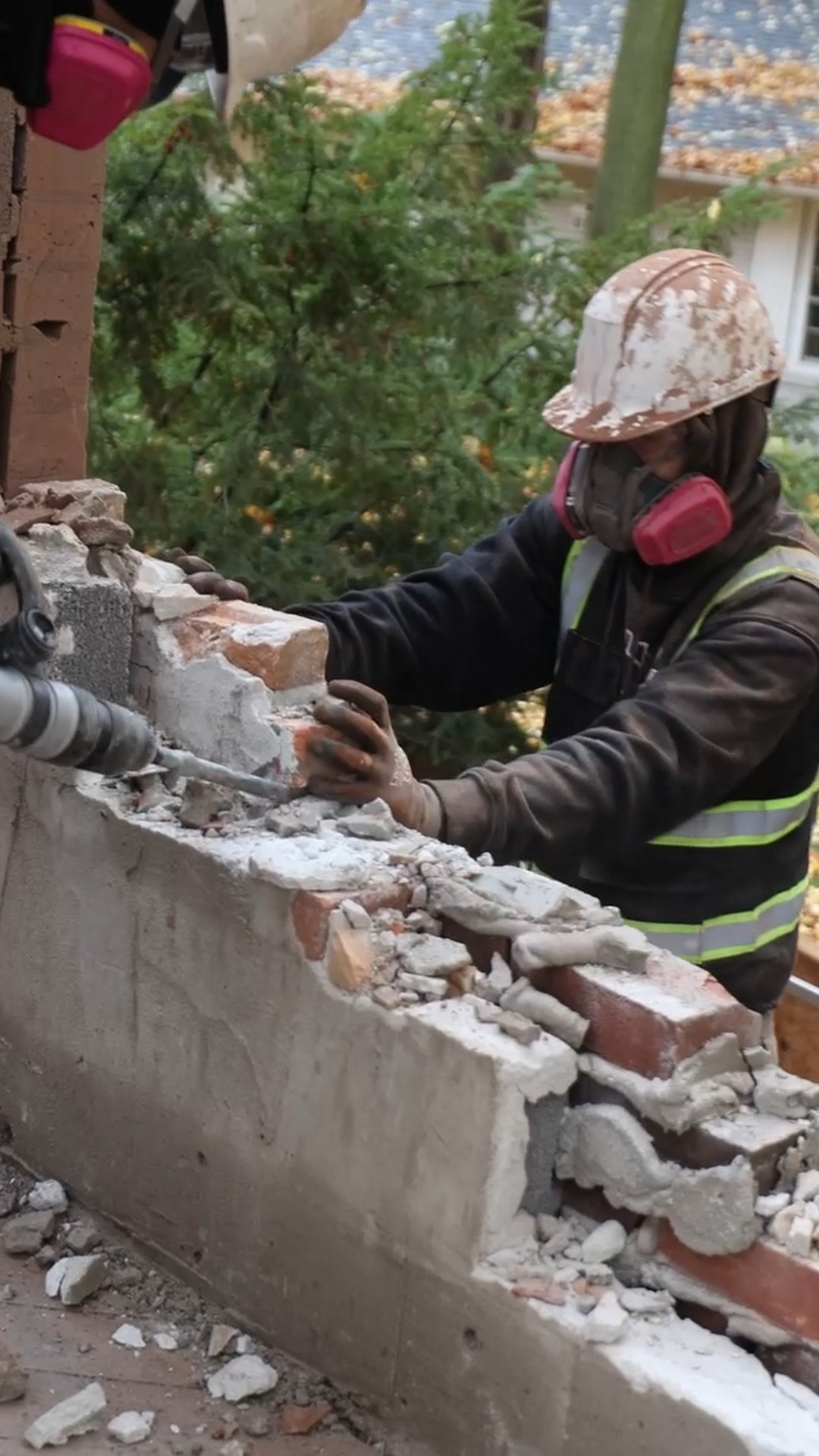 Worker in safety gear demolishing a brick and concrete wall with a power chisel.