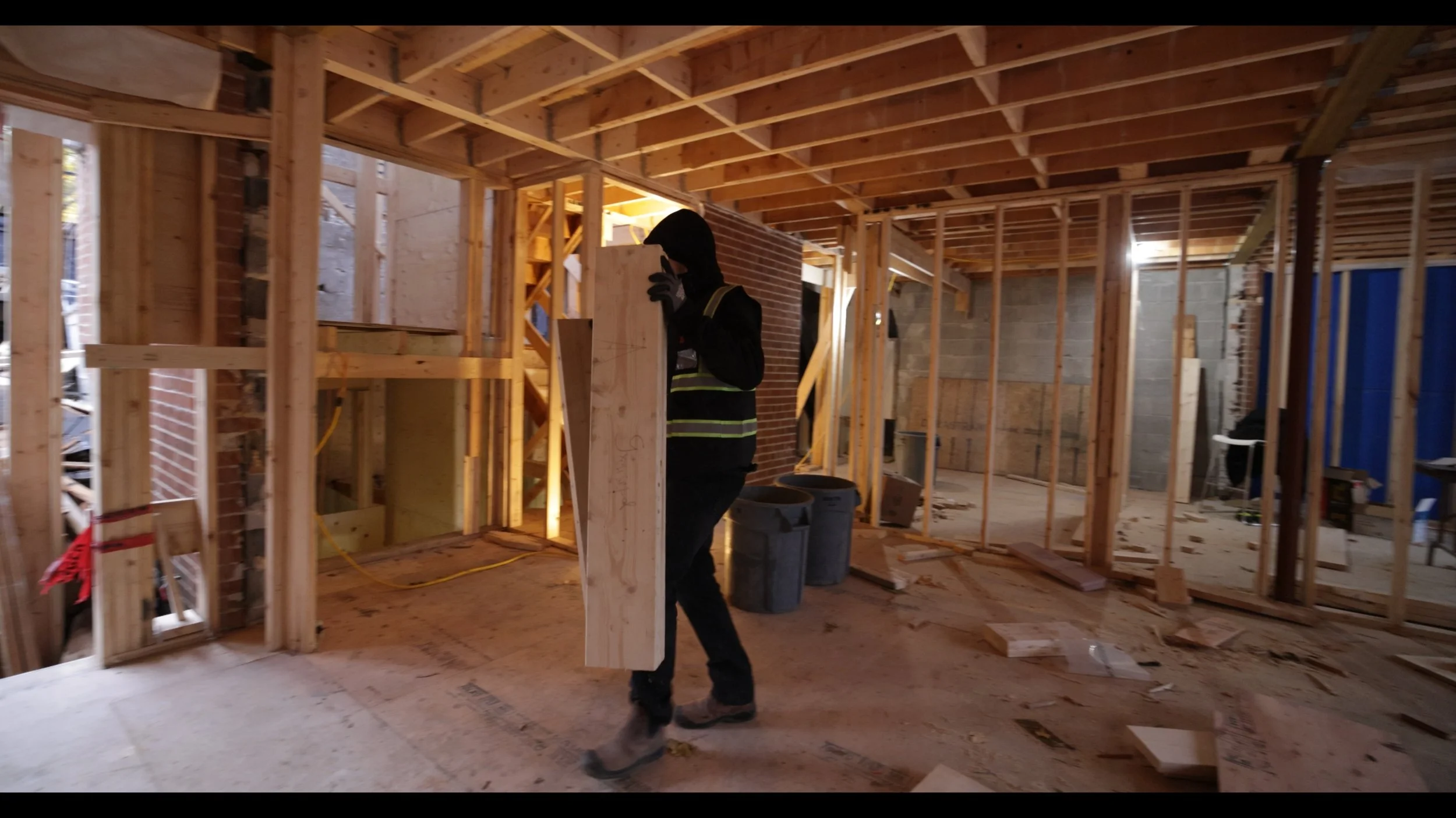 A construction worker carrying a wooden beam inside a building under construction with exposed wooden framing, brick, and concrete walls.