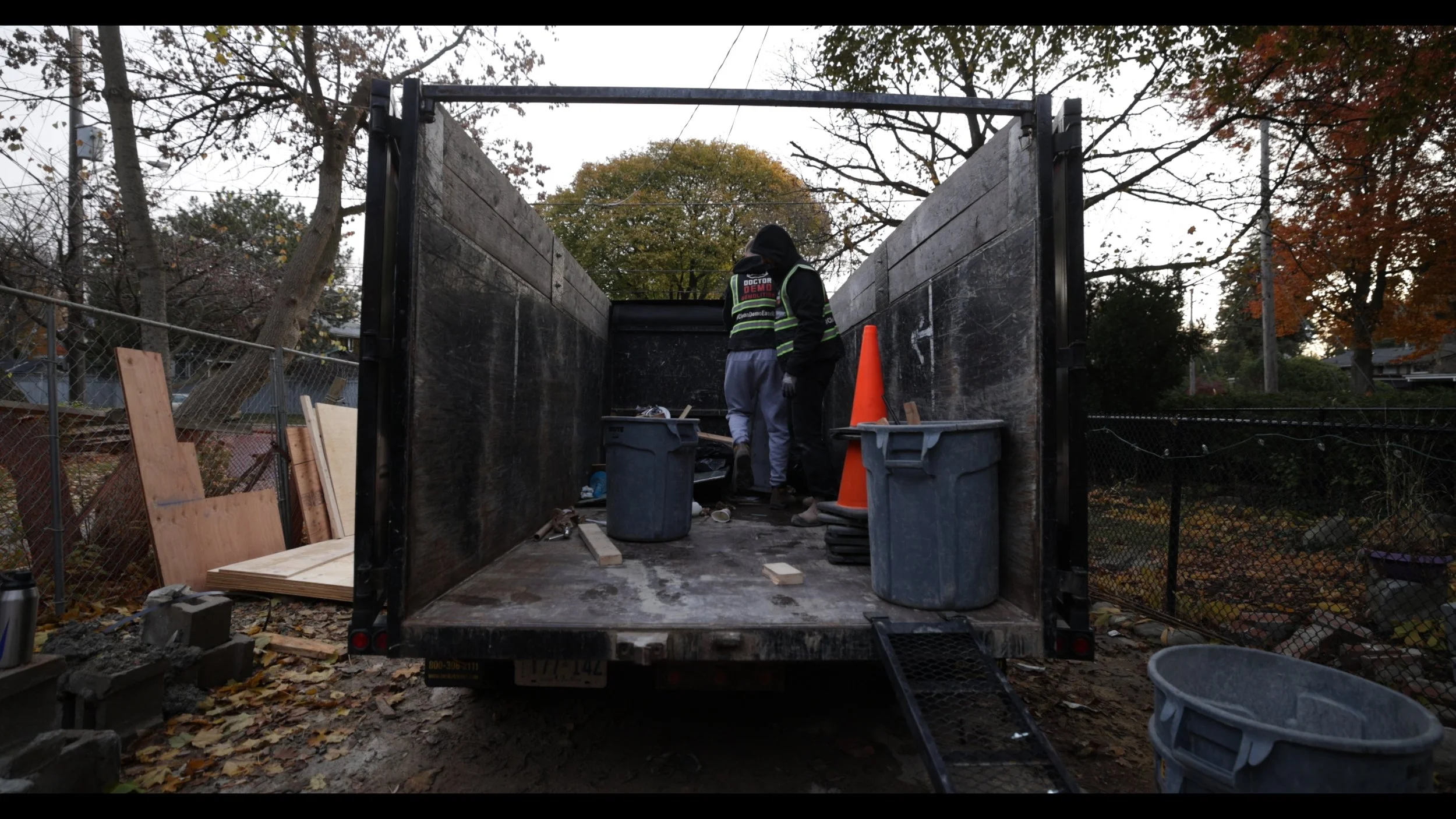 People working inside the back of a flatbed truck during daylight, with trees and a fence in the background, surrounded by construction materials and equipment.