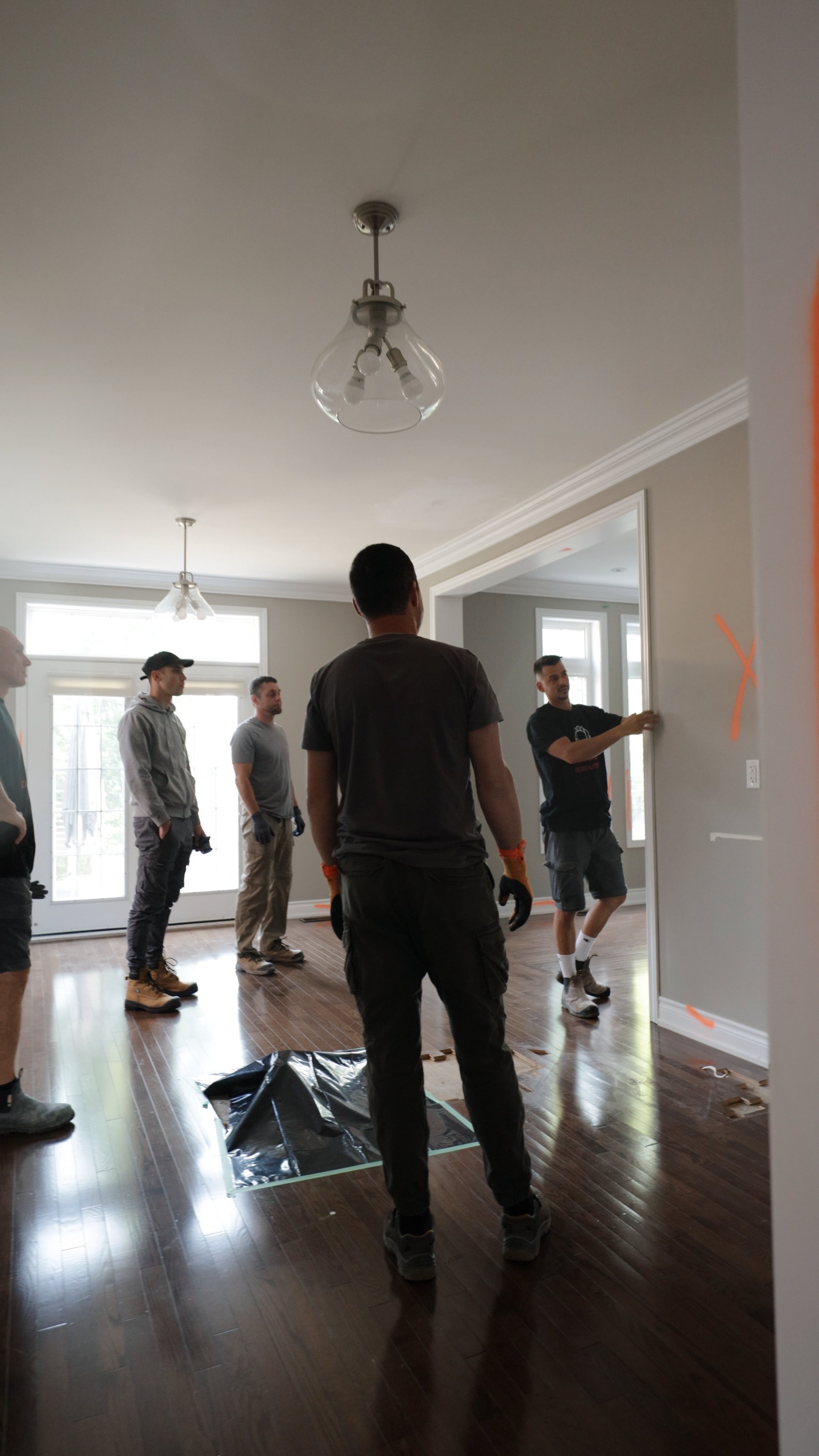 A group of five men standing inside a house, observing one of them pointing to a wall while others look on. The room has white walls, large windows, hardwood flooring, and modern ceiling light fixtures.