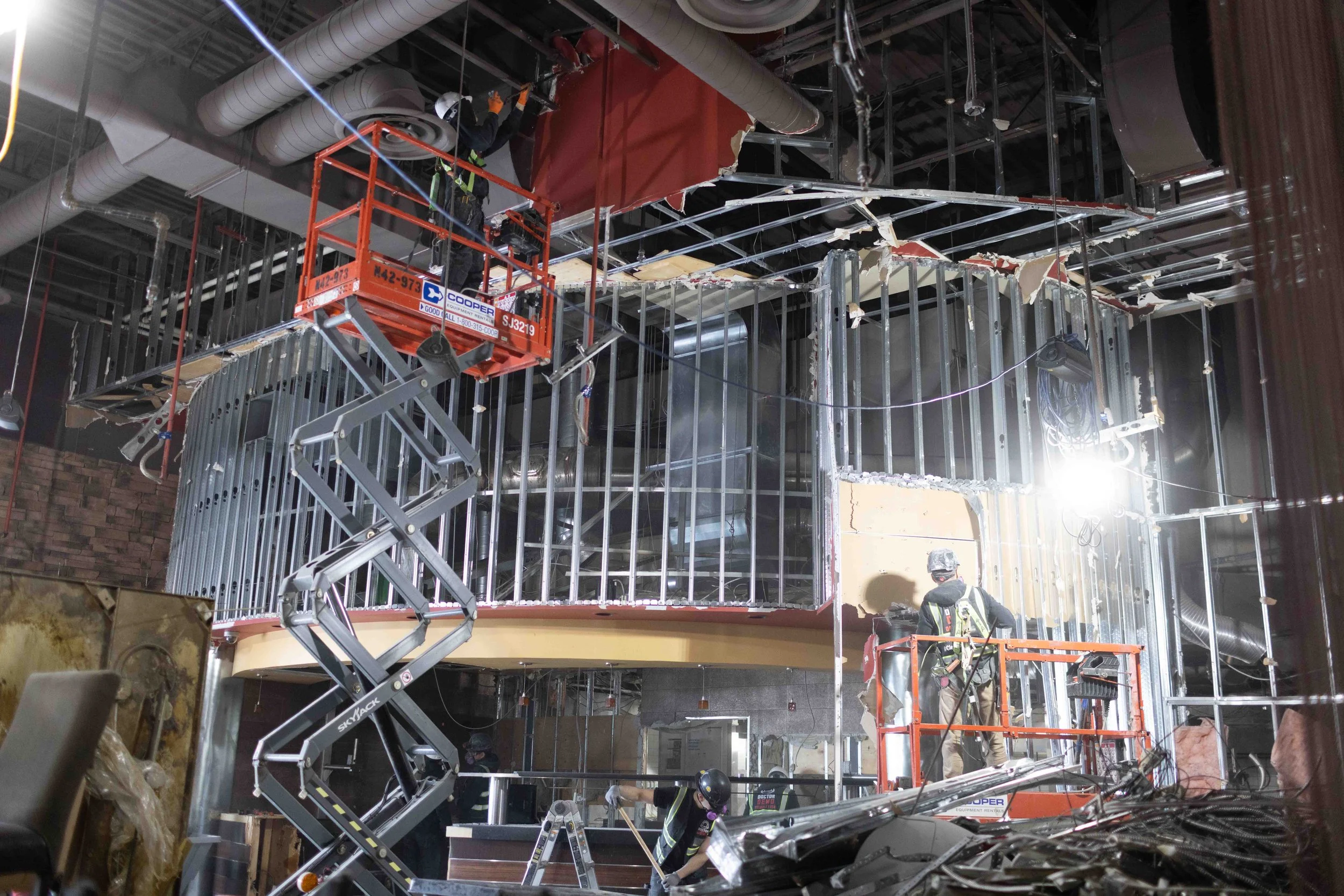 Construction workers installing ceiling panels in a building with exposed metal framing and ductwork, using scissor lifts.