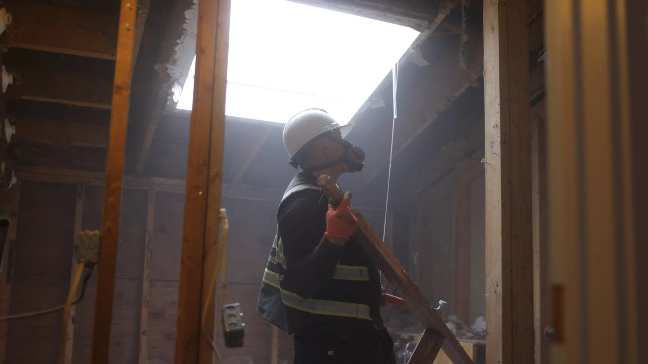 A construction worker wearing a hard hat and safety gloves inside a building under renovation, standing near an unfinished wall with visible wooden studs and a skylight overhead.