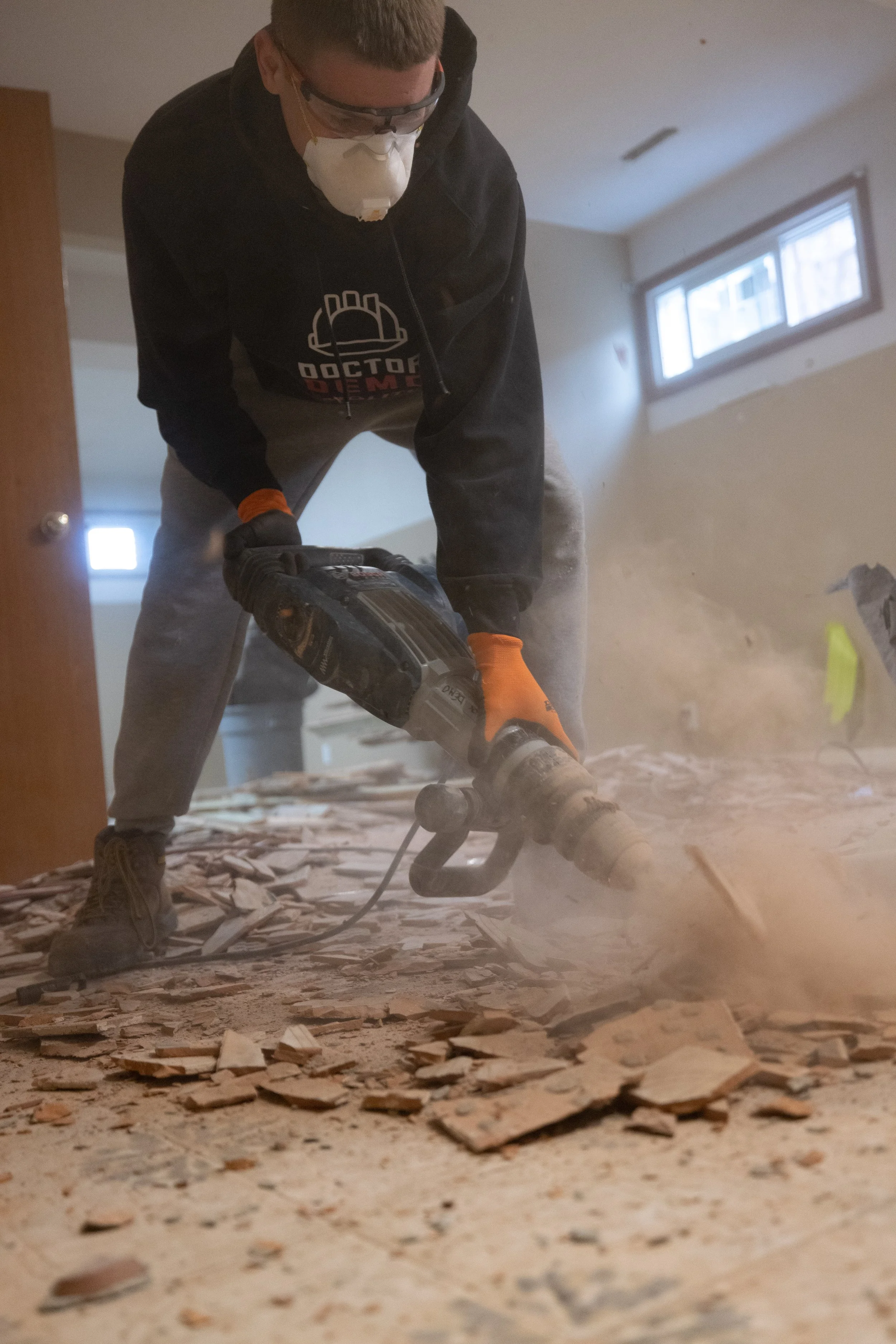 A person wearing a black hoodie, safety glasses, and a mask is using a power saw to cut through wood debris on a dusty floor in a room with a window and a door.