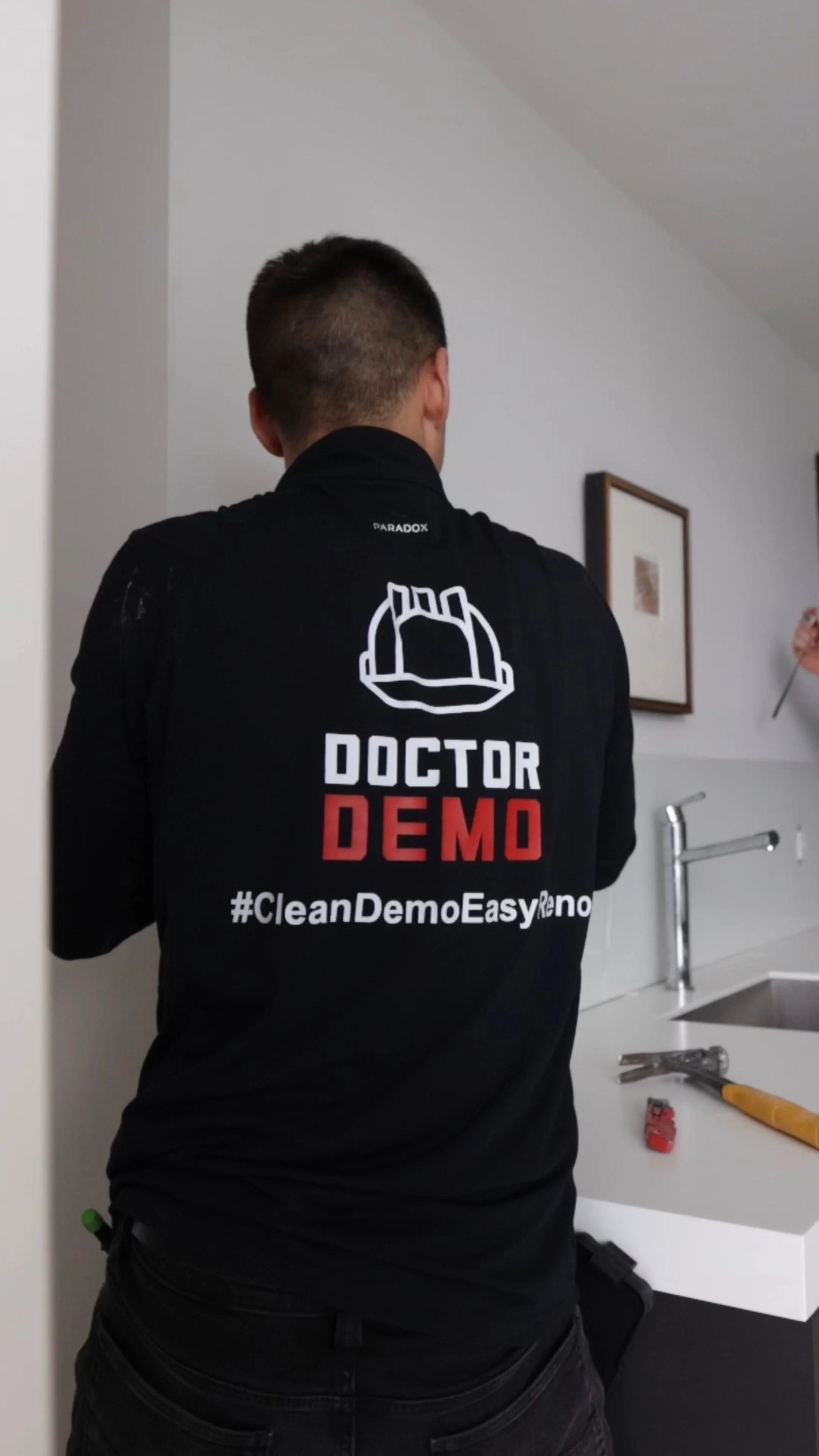 A man is standing in a kitchen wearing a black jacket with a logo of a hard hat and the text 'Doctor Demo' and '#CleanDemoEasyReno' on the back. The kitchen has a white countertop, a sink with a faucet, some tools on the counter, and framed pictures 