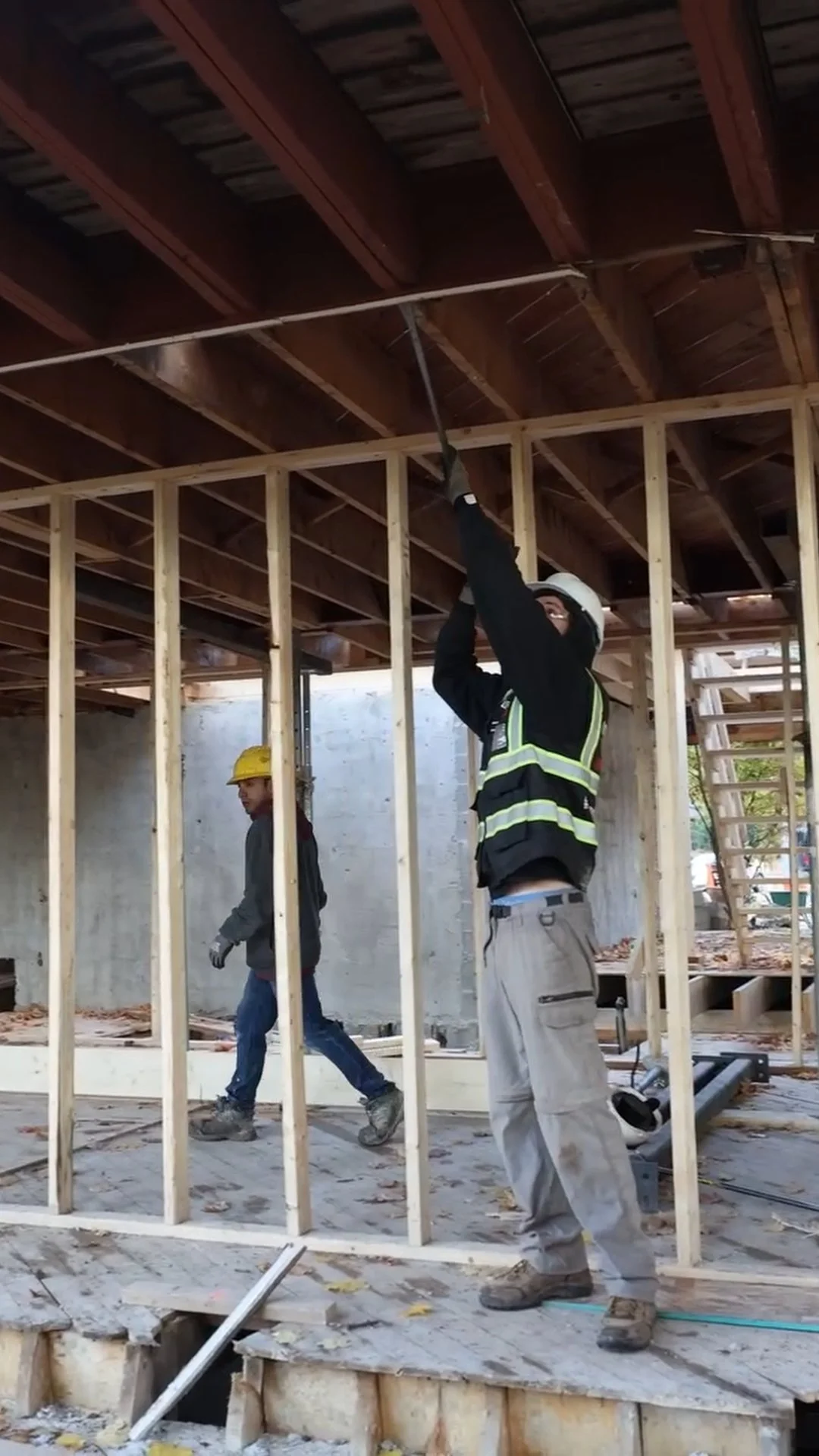 Two construction workers are working inside a building framed with wooden studs. One worker is using a tool on the ceiling, while the other walks through the area. Both are wearing hard hats and safety gear.
