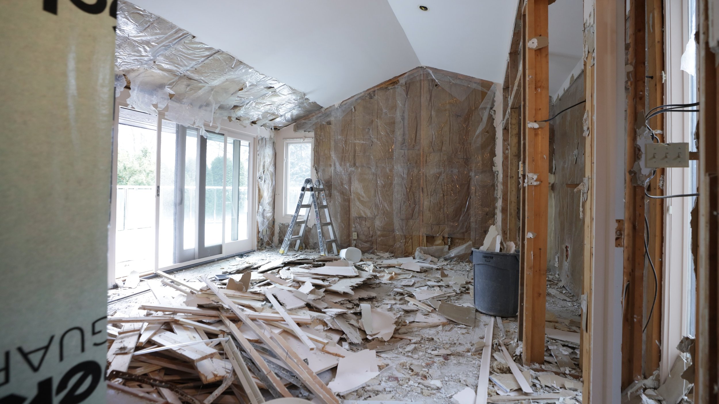 Room under renovation with debris on the floor, exposed wooden studs, a ladder, and ceiling insulation.