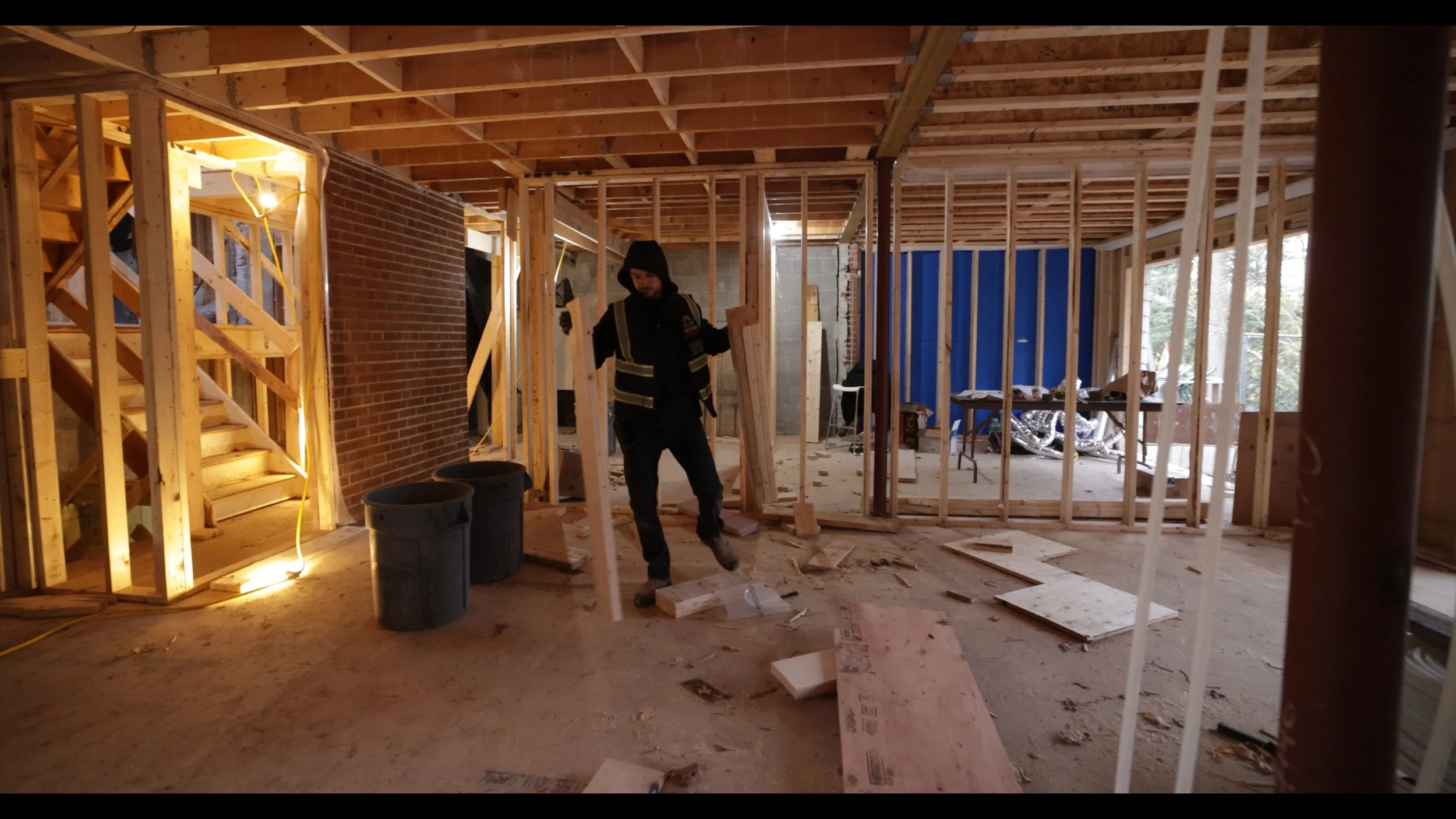 Construction worker in hoodie and safety vest walking inside a partly framed house under construction, with exposed wooden beams, wall studs, and construction materials scattered around.