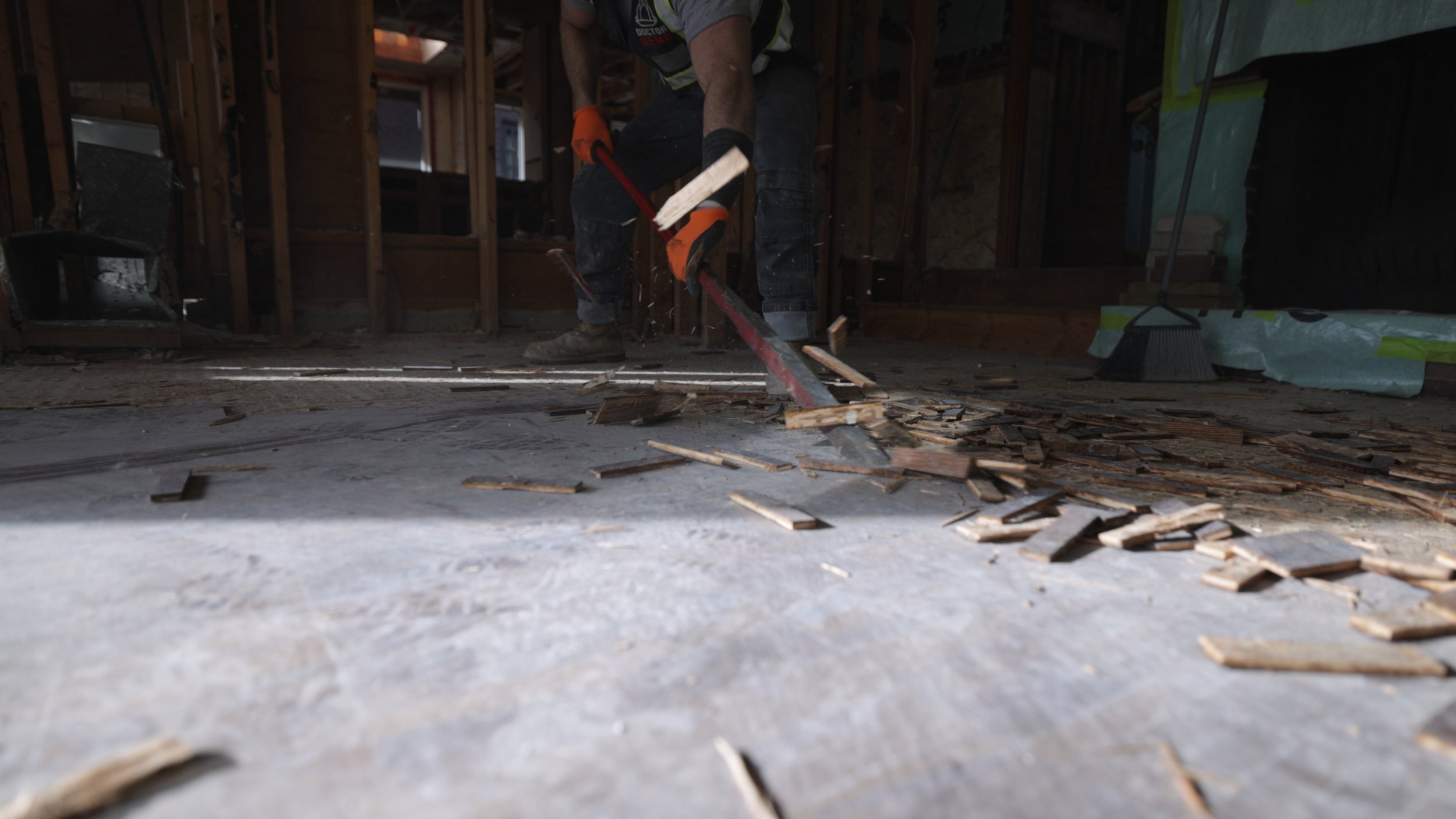 A person using a saw to cut wood inside a house under construction, with wood debris scattered on the floor.