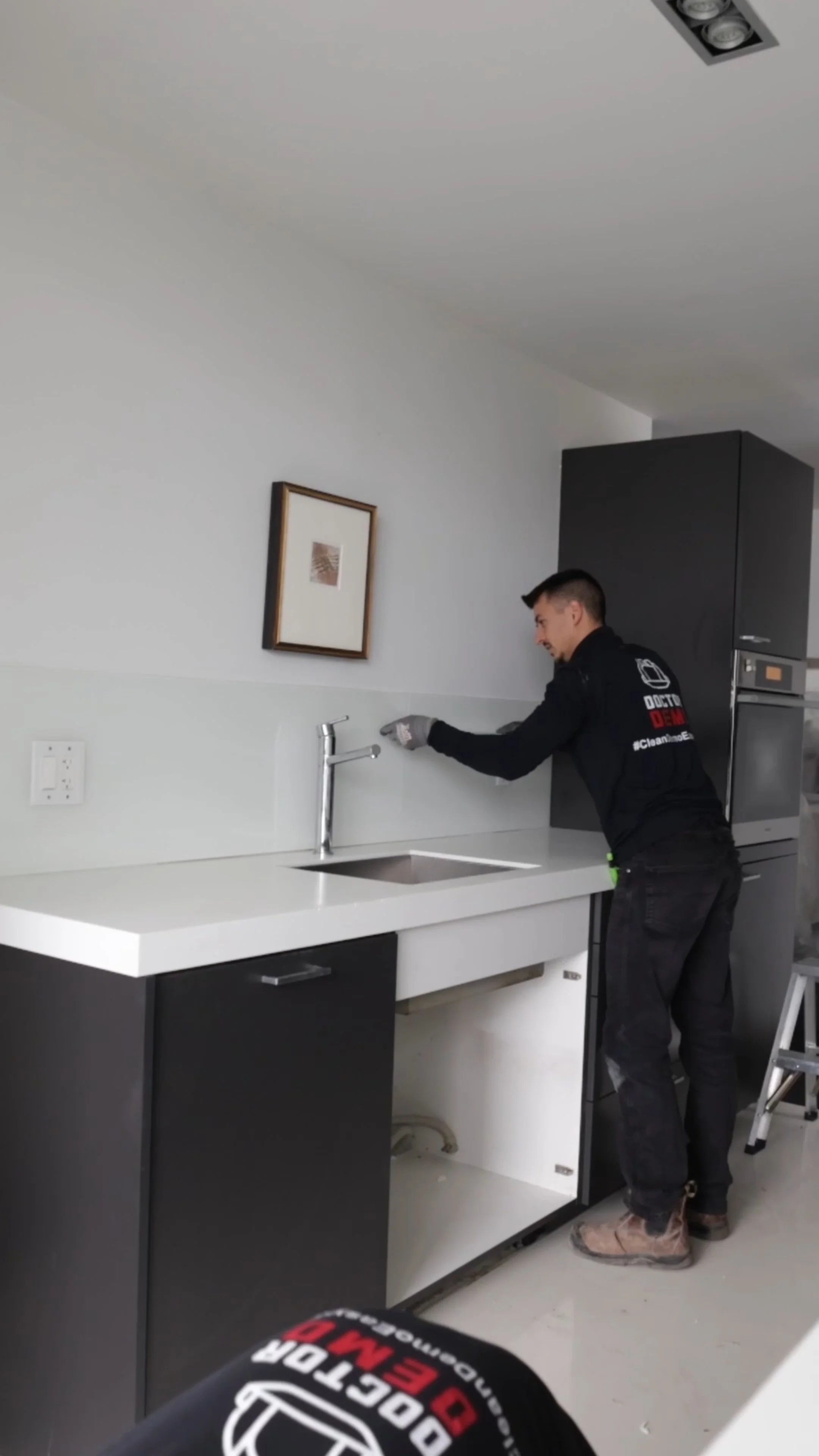 A technician installing or repairing a kitchen cabinet under a white countertop. The technician is wearing a black uniform with a logo that says 'Door & Window' and is standing on a small ladder.