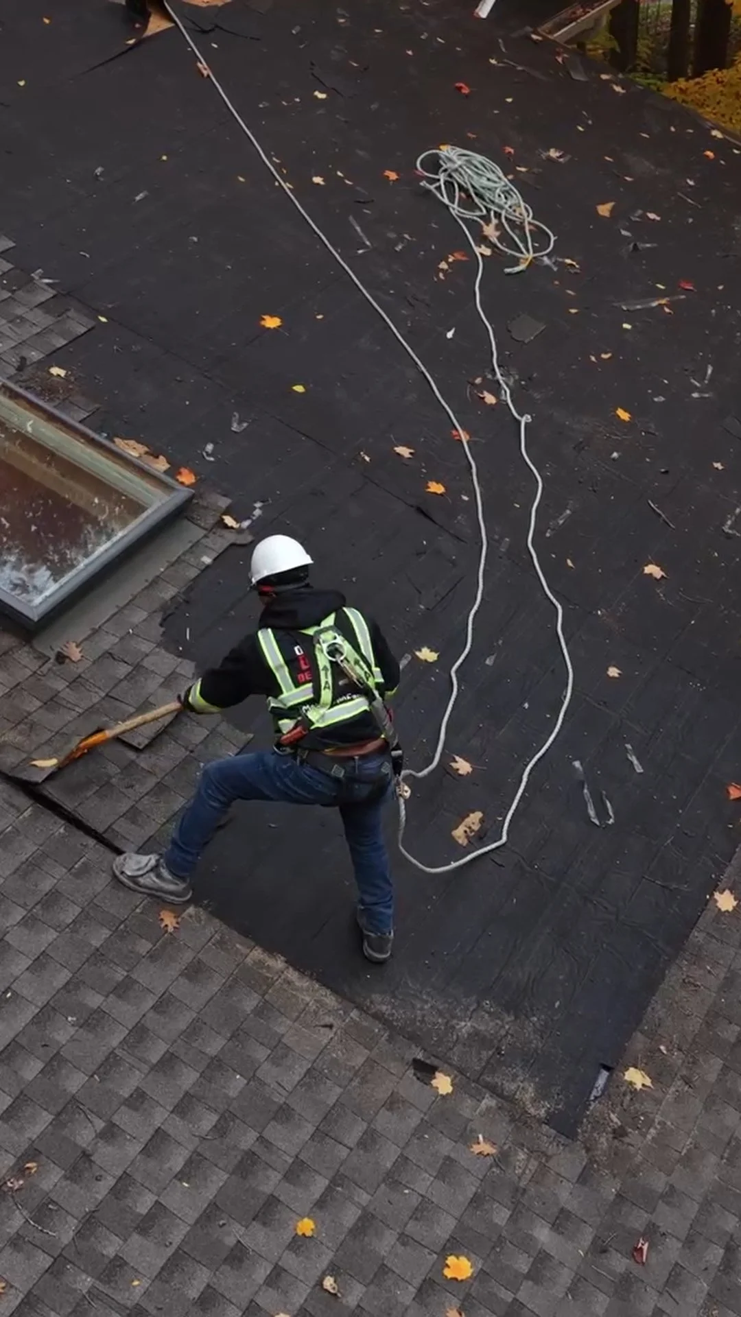 A person wearing a hard hat, safety vest, and jeans is using a rake to clear debris from a roof. A coil of white rope is on the roof, and autumn leaves are scattered across the surface.