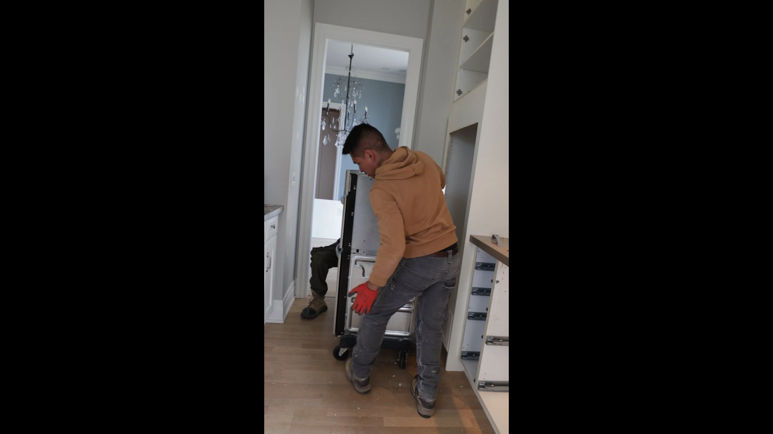 Two men are moving a black appliance through a doorway in a home. One man is wearing a tan hoodie and gray jeans, the other is in a black jacket and dark pants.