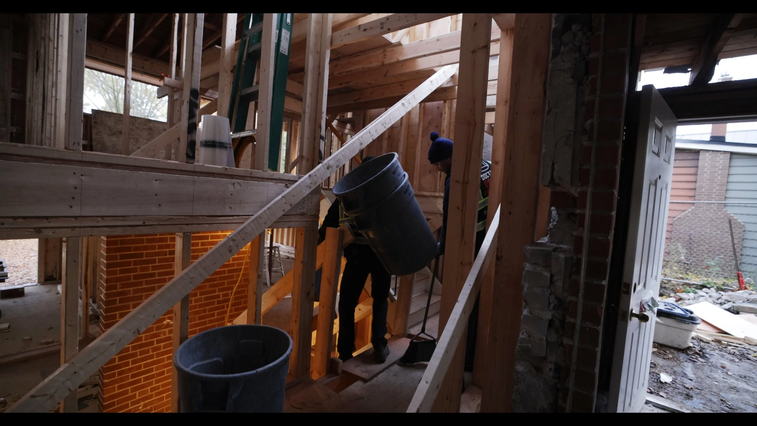 Construction workers building a wooden staircase inside a house under construction, with exposed wooden framing and an open door leading outside.