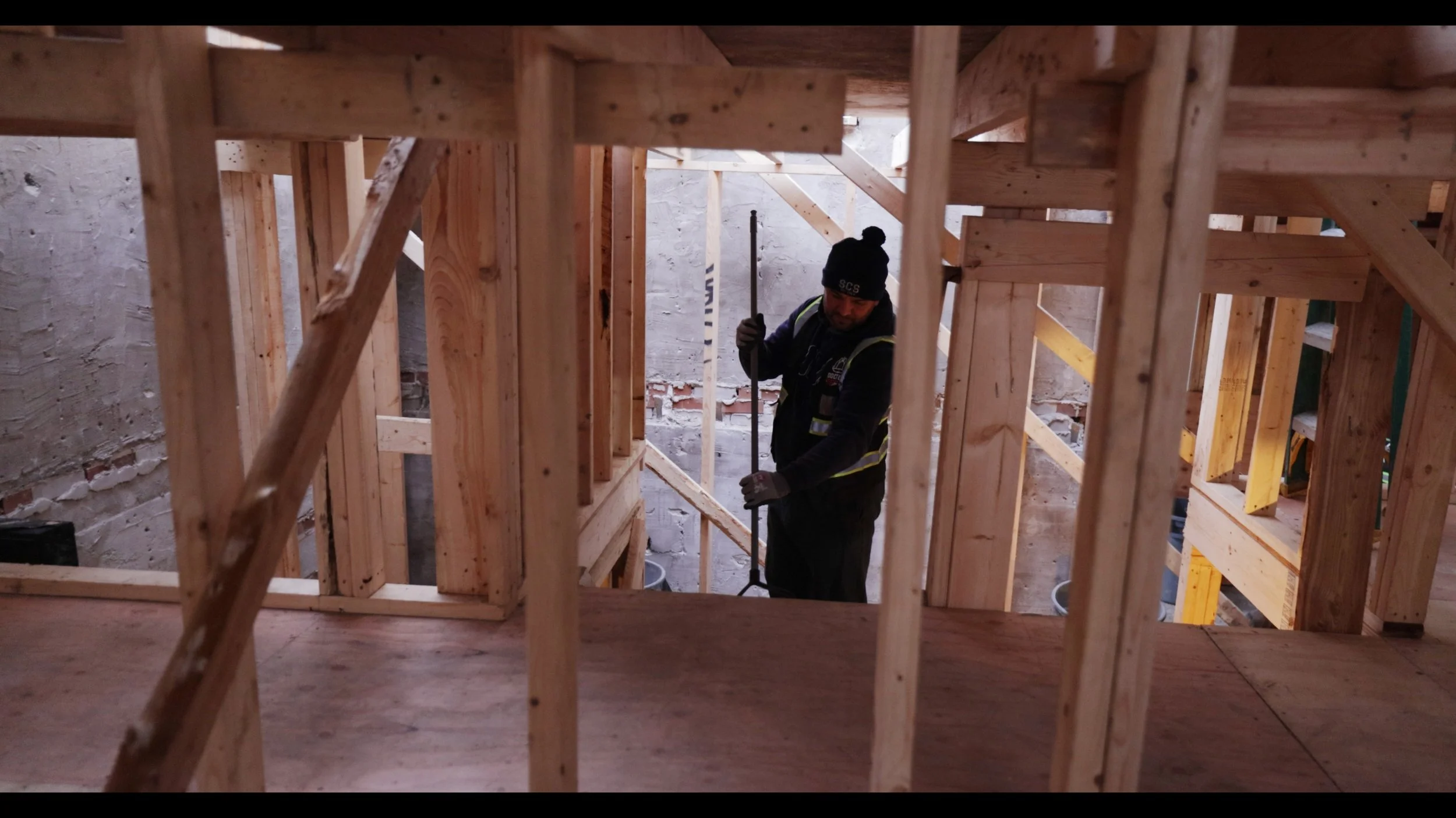 Construction worker inside a wooden frame structure at a building site, holding a tool.