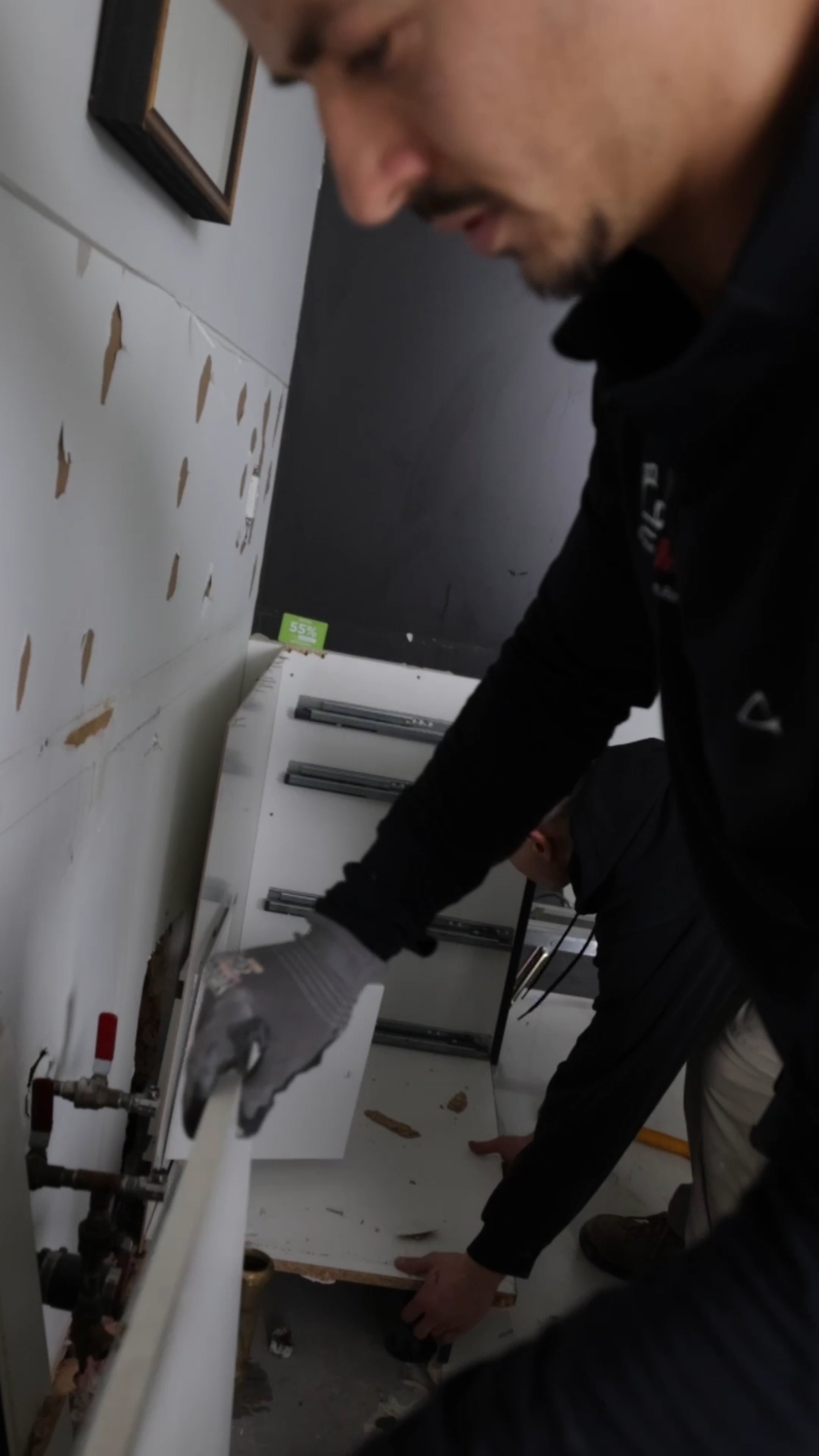 Two maintenance workers working on a wall-mounted unit with peeling paint, wearing gloves and safety gear in an indoor setting.