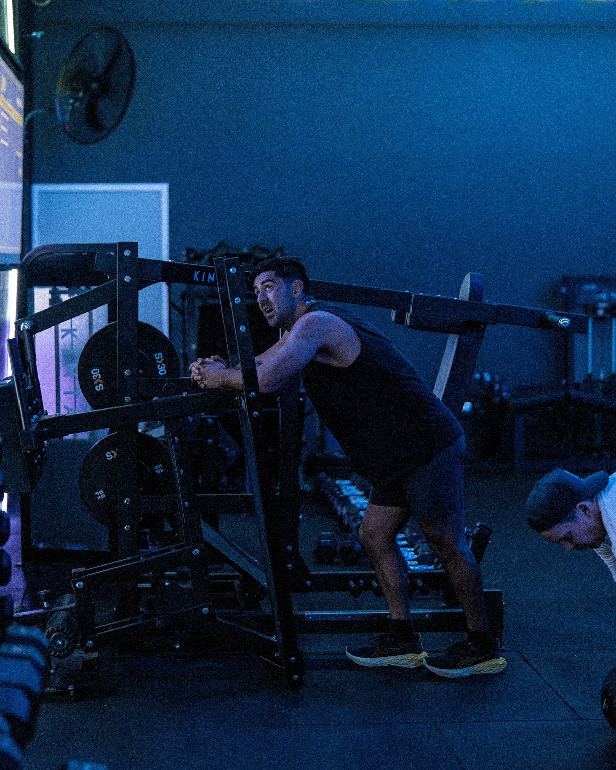 Black and white photo of a gym with various exercise equipment, kettlebells, and weights, with large windows in the background. Overlaid text in blue reads "OPEN GYM 24/7."