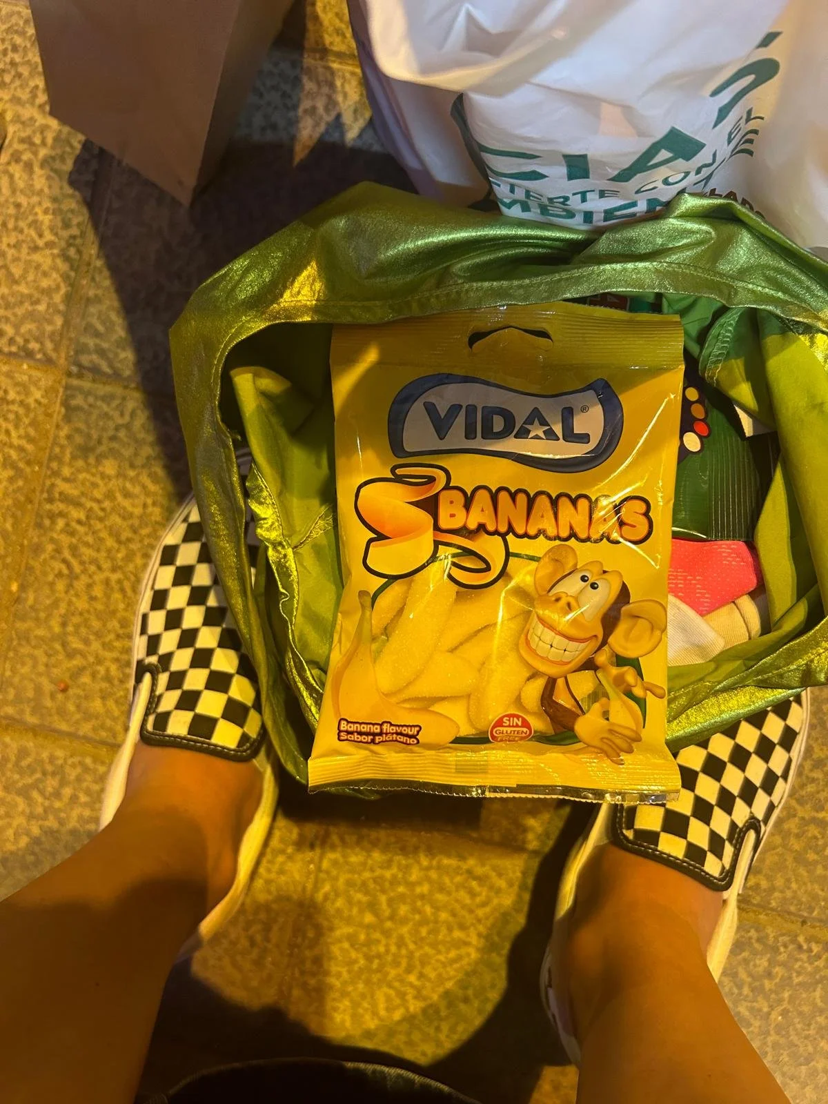 Tasche mit einer Packung Bananen-Gummibärchen der Marke Vidal, abgesehen davon sind in der Tasche weitere Gegenstände, die nicht sichtbar sind, sowie Schuhe mit einem schwarz-weißen Checker-Design. Der Blick ist von oben auf die Schuhe und die Tasche gerichtet.