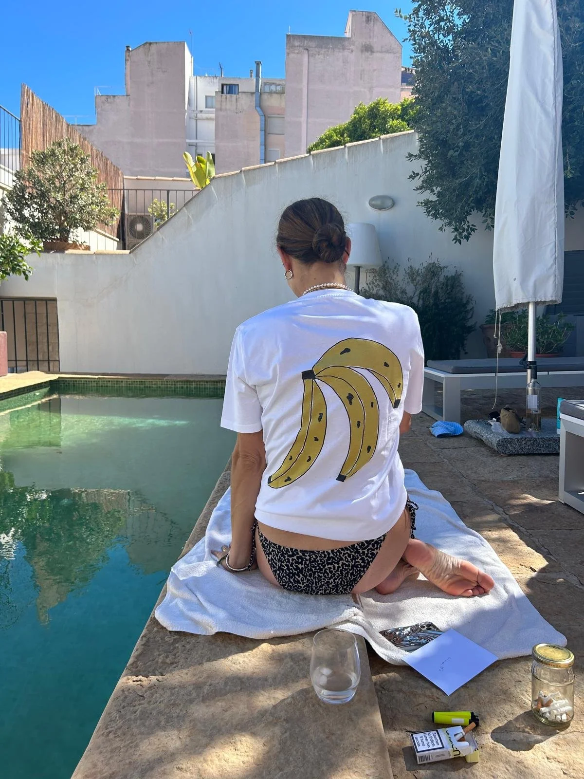Frau, die auf einem Tuch neben einem Pool sitzt, trägt ein weißes T-Shirt mit einer Banane auf der Rückseite, und trägt Leoparden-Boxershorts. Der Hintergrund zeigt einen weißen Mauern und einen blauen Himmel.