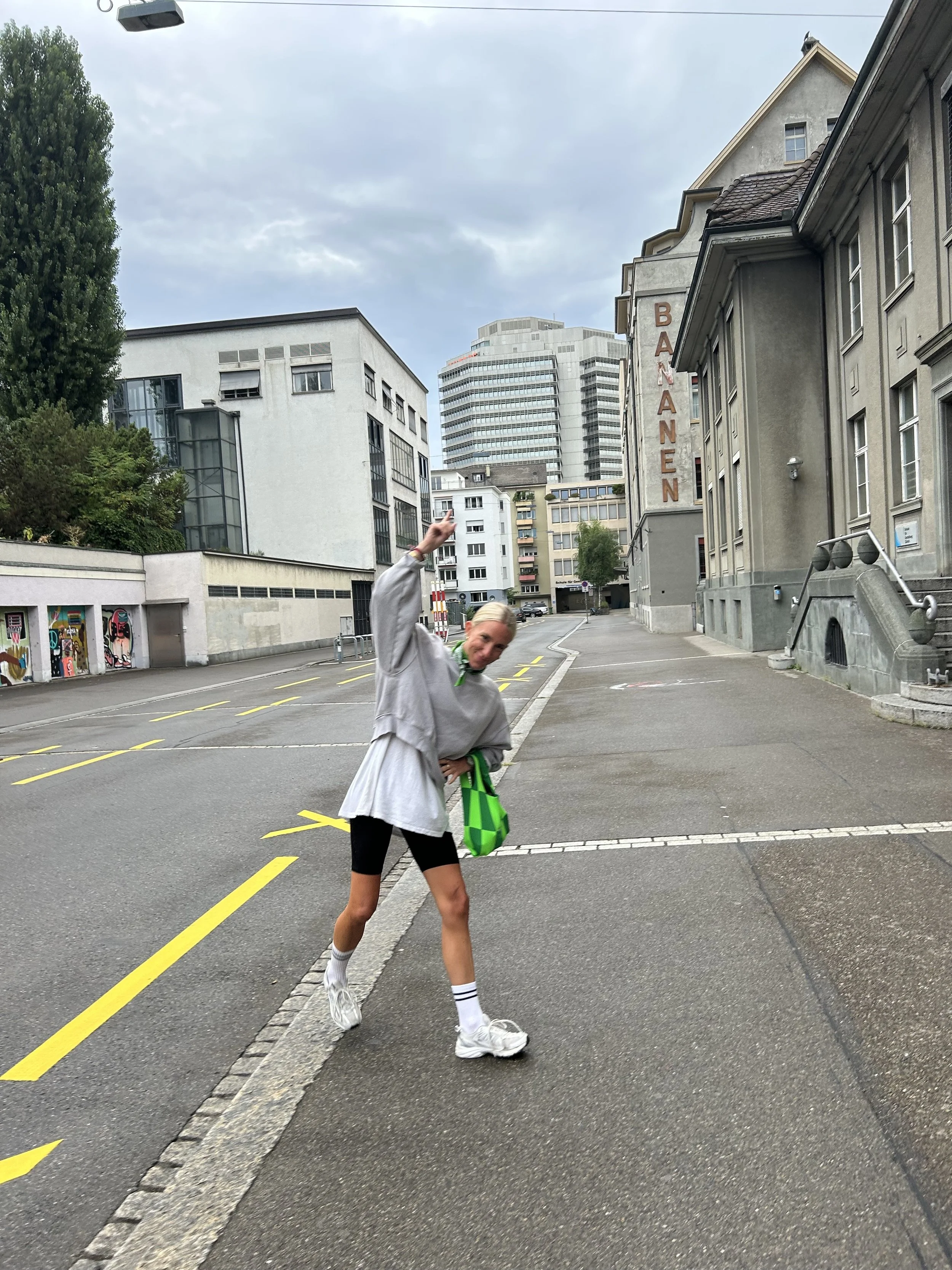 Eine junge Frau steht an einer Straßenkreuzung, trägt sportliche Kleidung und hält eine grün-weiße Tasche. Sie hebt den rechten Arm und lächelt, während sie vor einer urbanen Hintergrundkulisse mit Gebäuden steht.