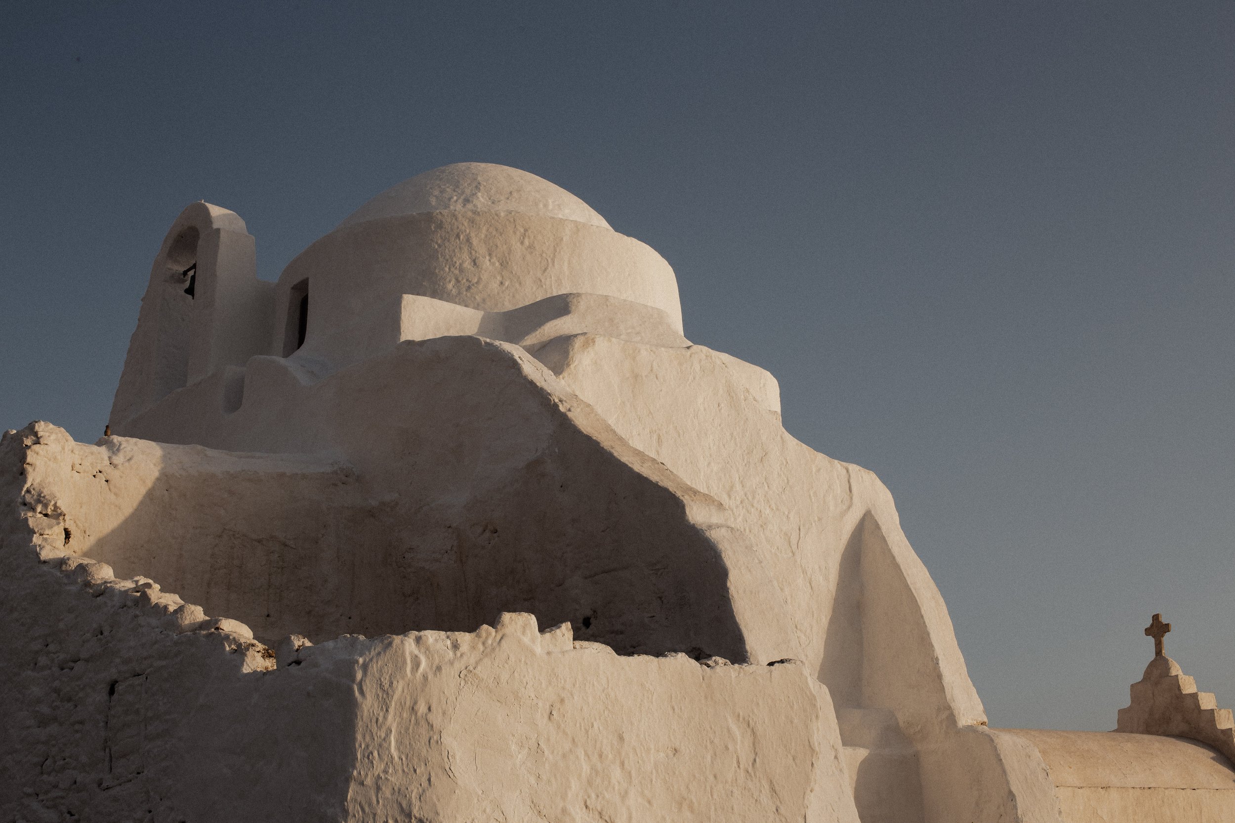 Whitewashed church with a dome and a bell tower against a clear sky, featuring a cross on a nearby structure.